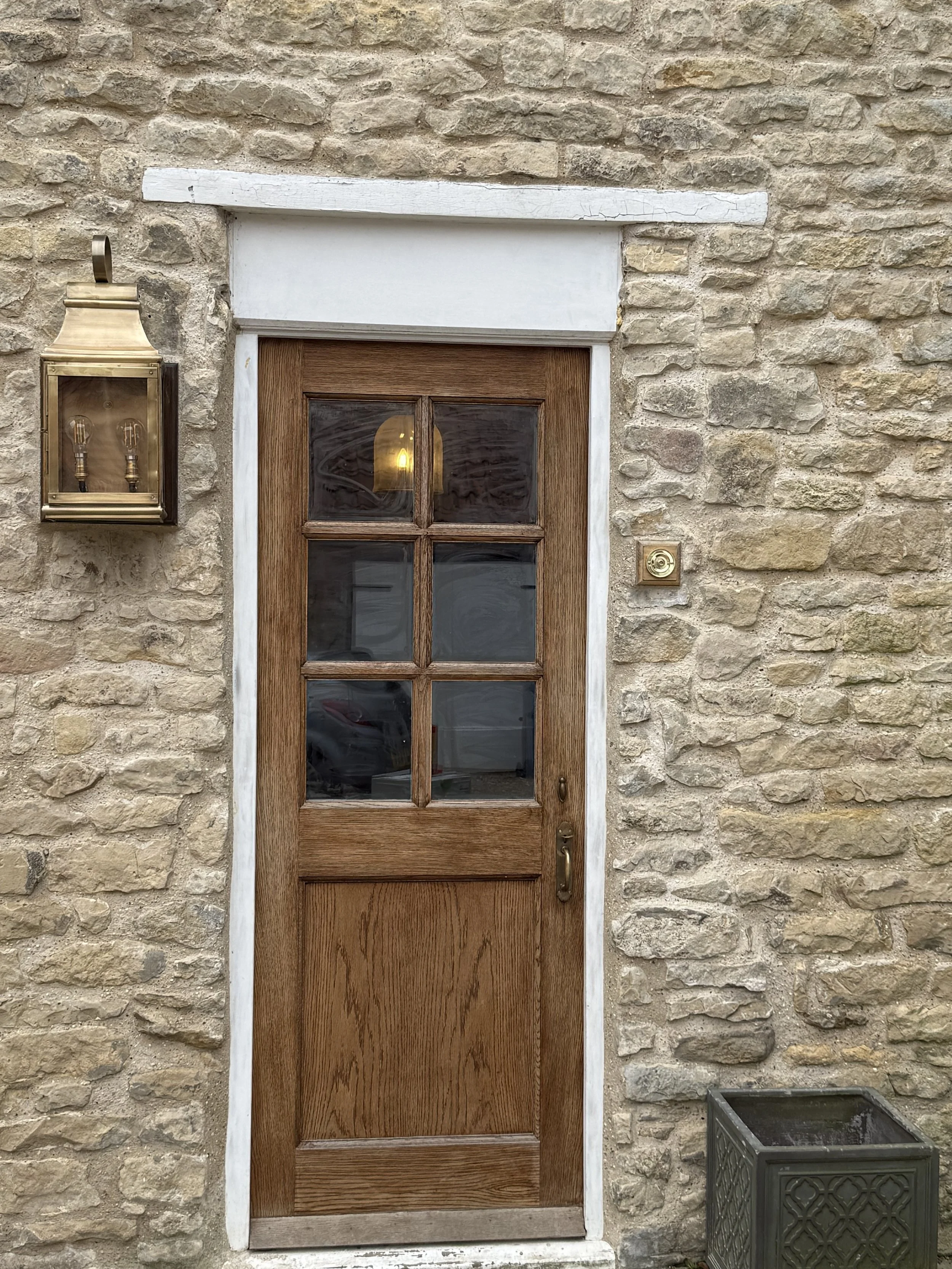Wooden front door with six glass panes on a stone wall, accompanied by an outdoor light fixture and a decorative planter.