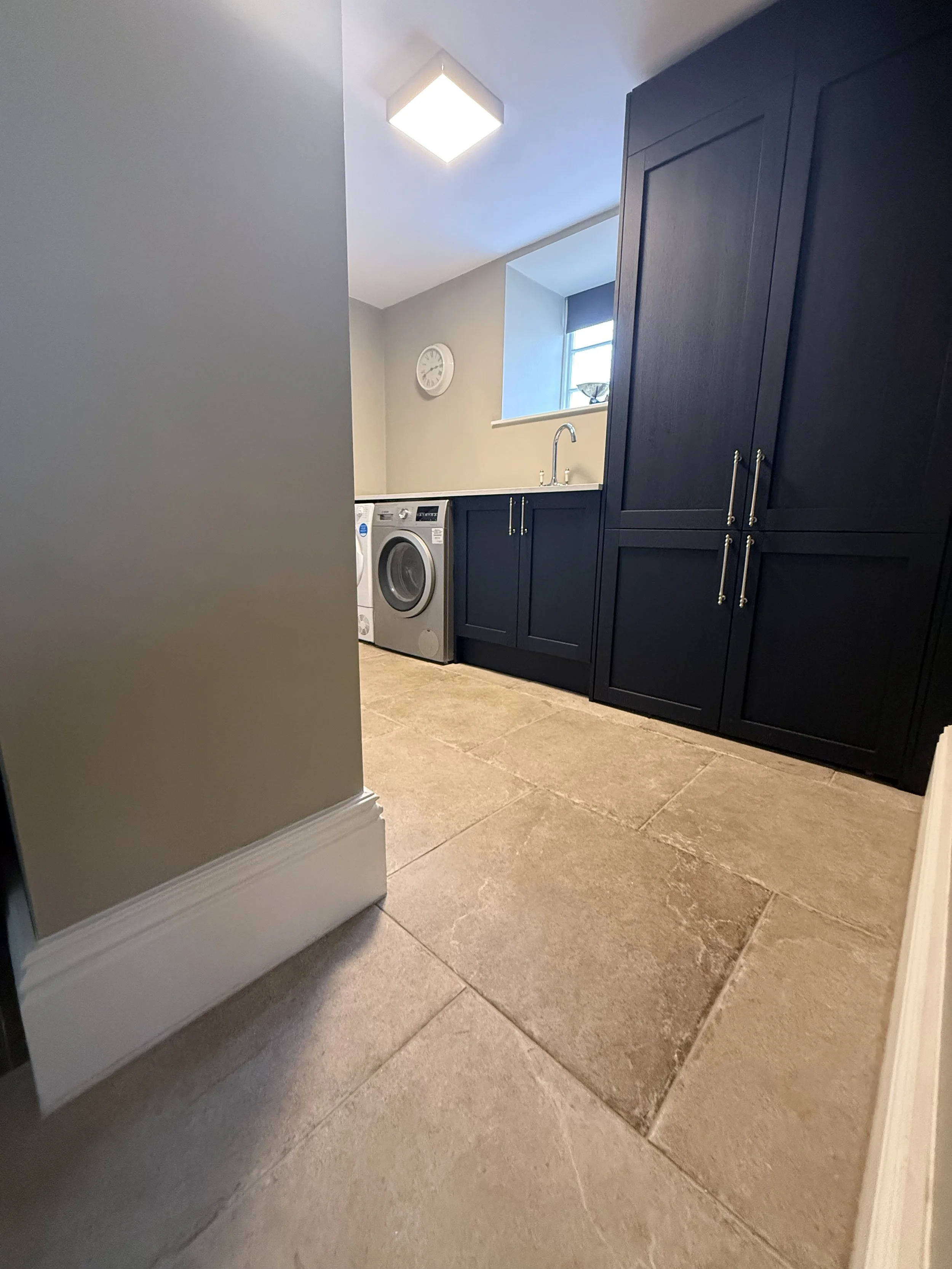 A laundry room with beige tiled floor, beige walls, dark blue cabinets, a small window, a washing machine, a sink, and a wall clock.