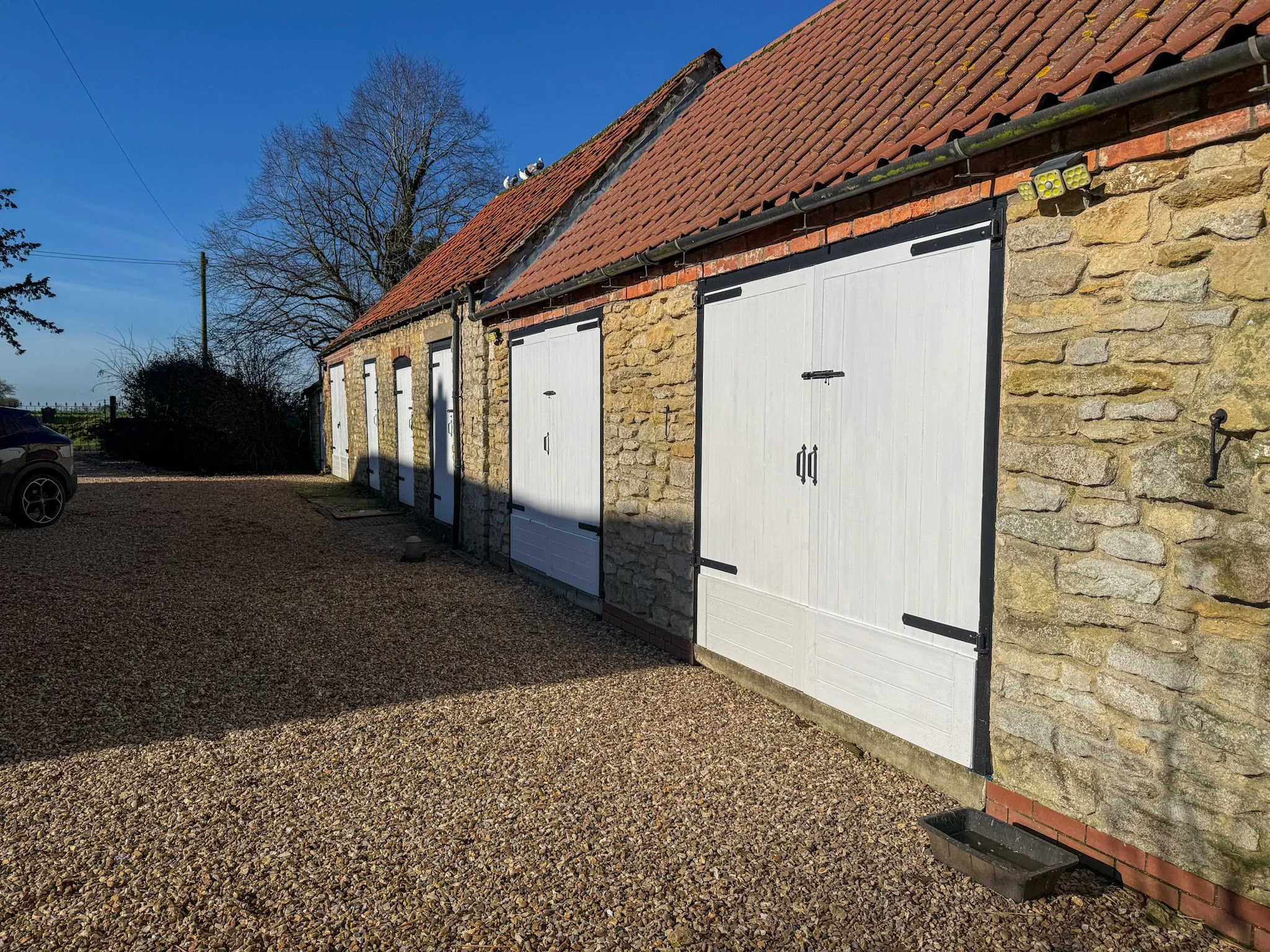 A row of stone garages with white doors and red-tiled roofs, a gravel driveway, and a black car parked on the left, under a clear blue sky with a leafless tree in the background.
