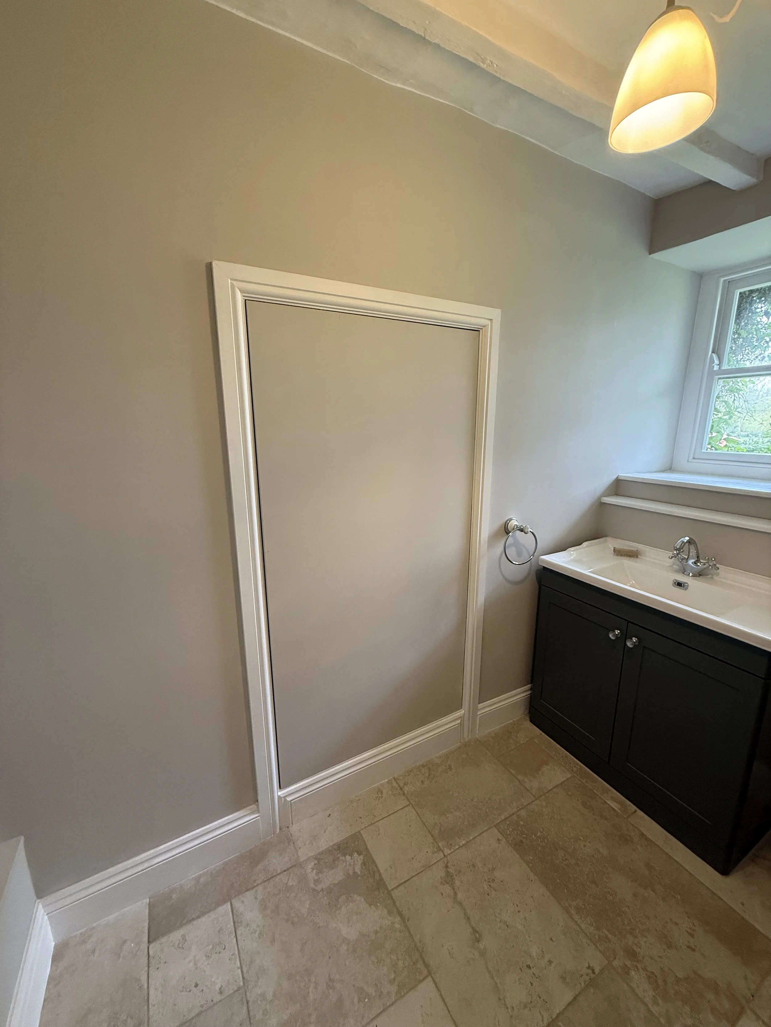 A bathroom with beige walls, a small door, a dark wooden vanity with a white sink, a window with a view of greenery, and a ceiling light.