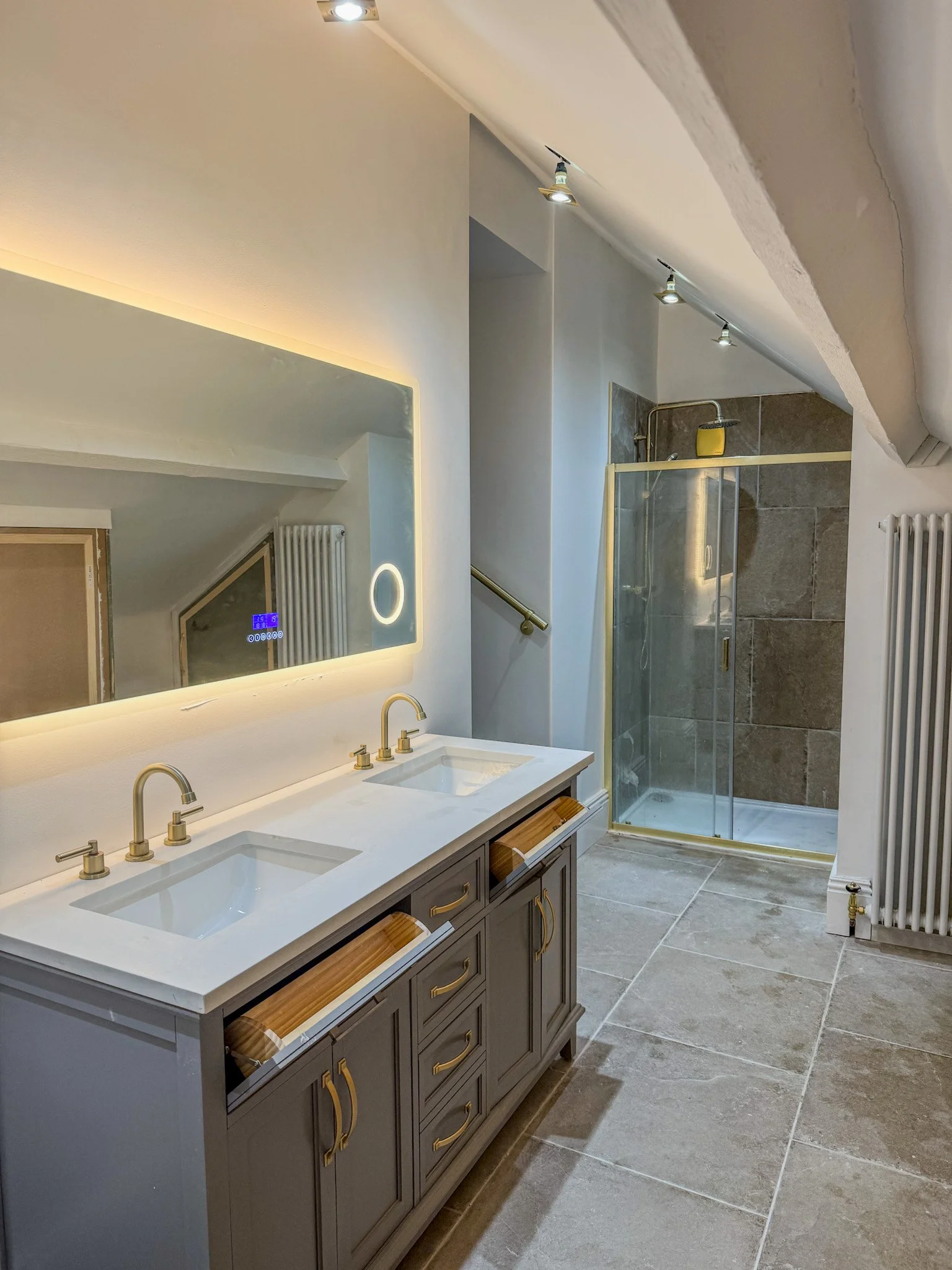 A modern bathroom with a double vanity sink with a large illuminated mirror, a glass shower enclosure with brown tile walls, a white radiator, and a sloped ceiling with spotlights.