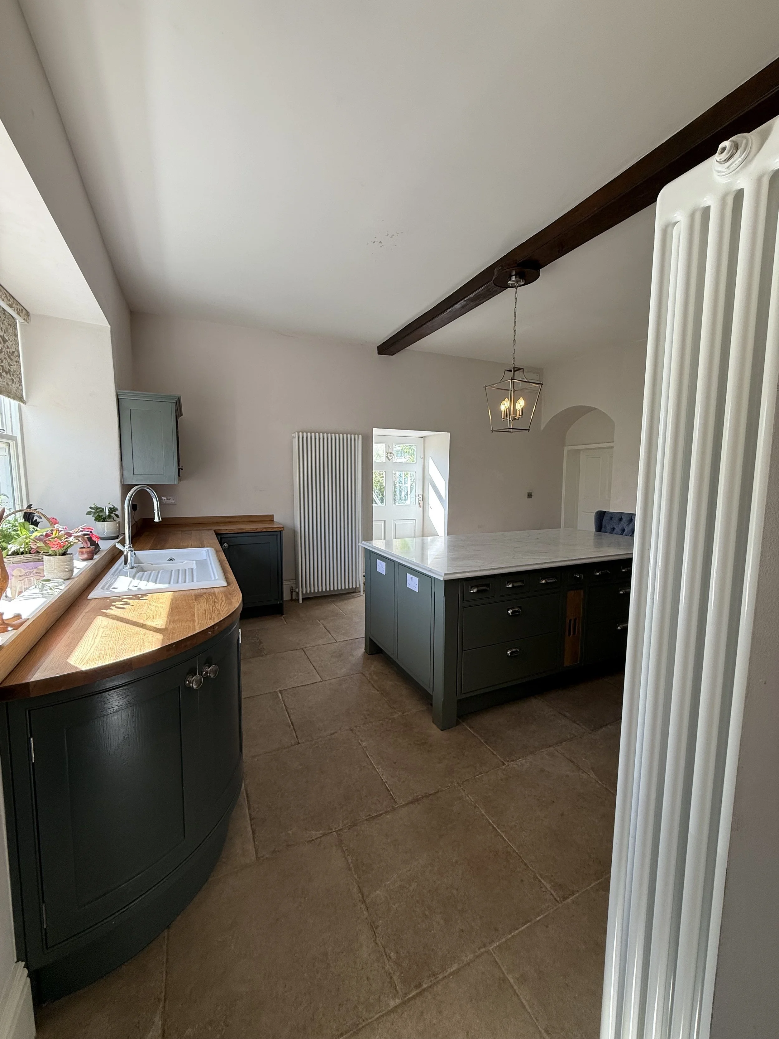 A spacious kitchen with a curved wooden countertop, blue cabinets, a marble island, large window with potted plants, and a door leading outside. Neutral-colored floor tiles and a hanging light fixture are visible.