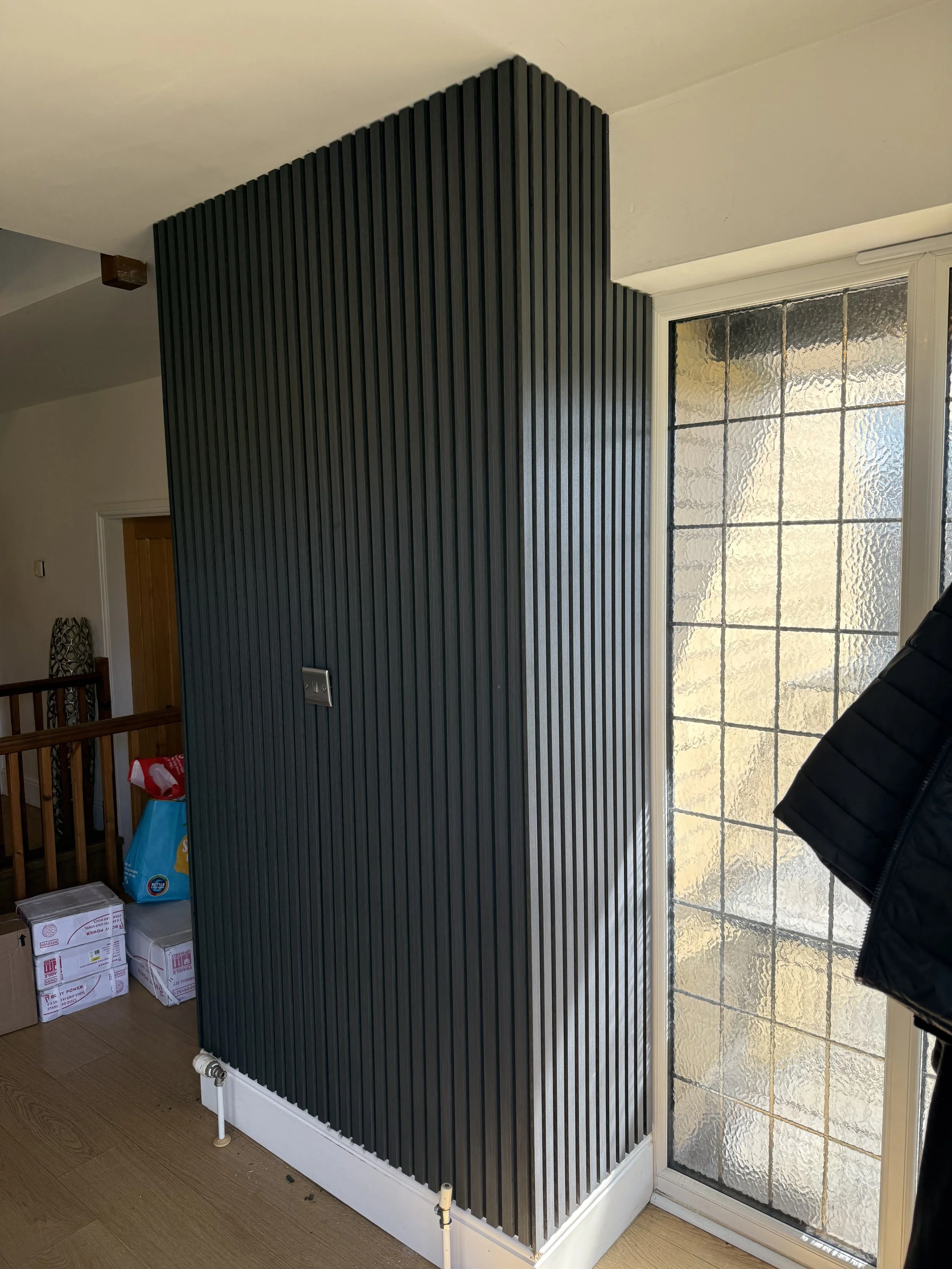 Interior view of a room with a black slatted wall panel next to a large textured glass window, wooden flooring, and some boxes and household items.