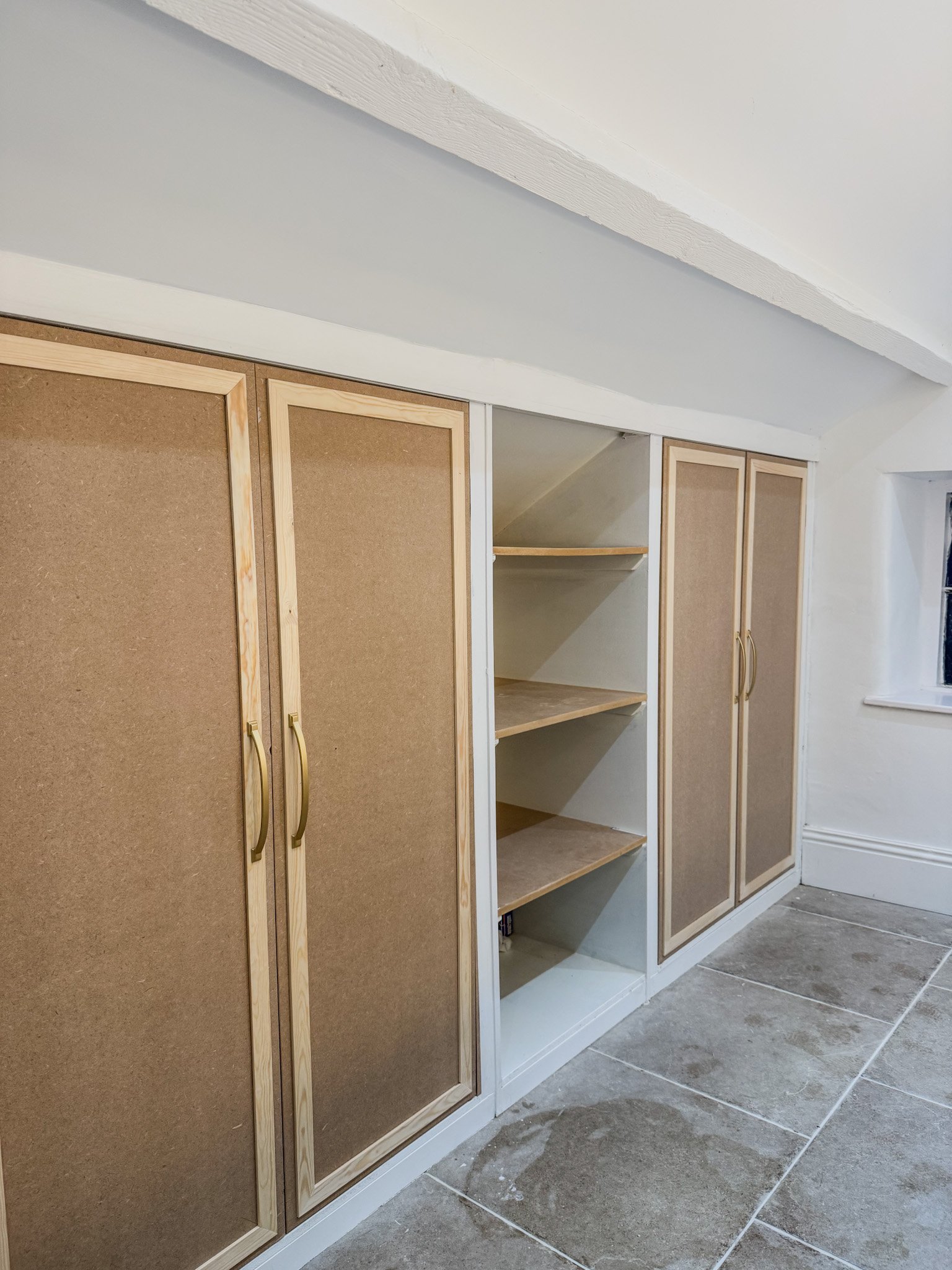 Construction of built-in shelves and cabinets in a room with a sloped ceiling, showing unfinished wood and tan insulated panels.