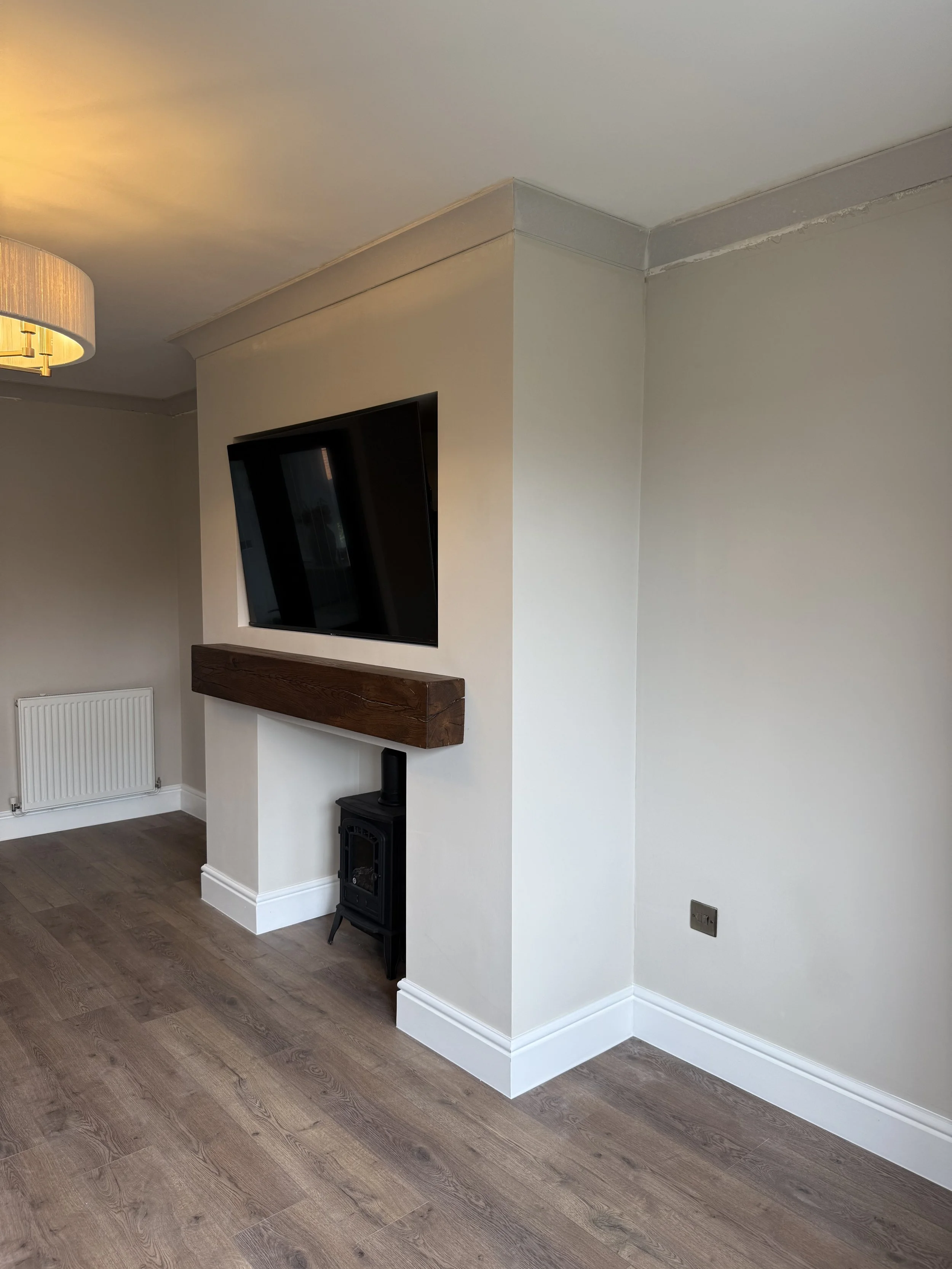 A corner of a room with a flat-screen TV mounted on the wall above a wooden shelf. Below the shelf, there is a small black stove on the floor. The floor is wood, and the walls are painted white. There is a radiator on the left wall and a power outlet