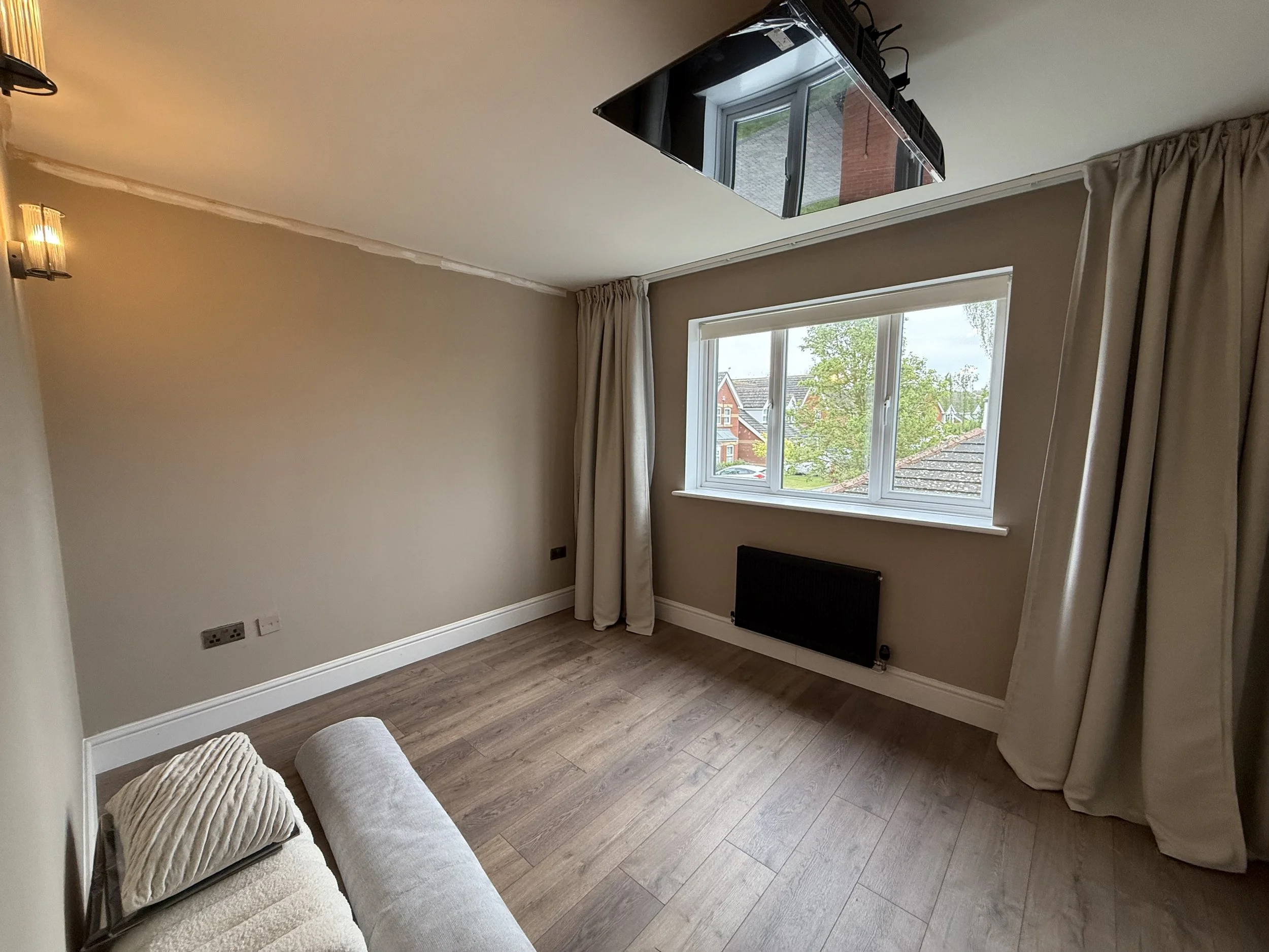 Living room with a window showing houses outside, beige walls, curtains, a ceiling light, and a gray ottoman.
