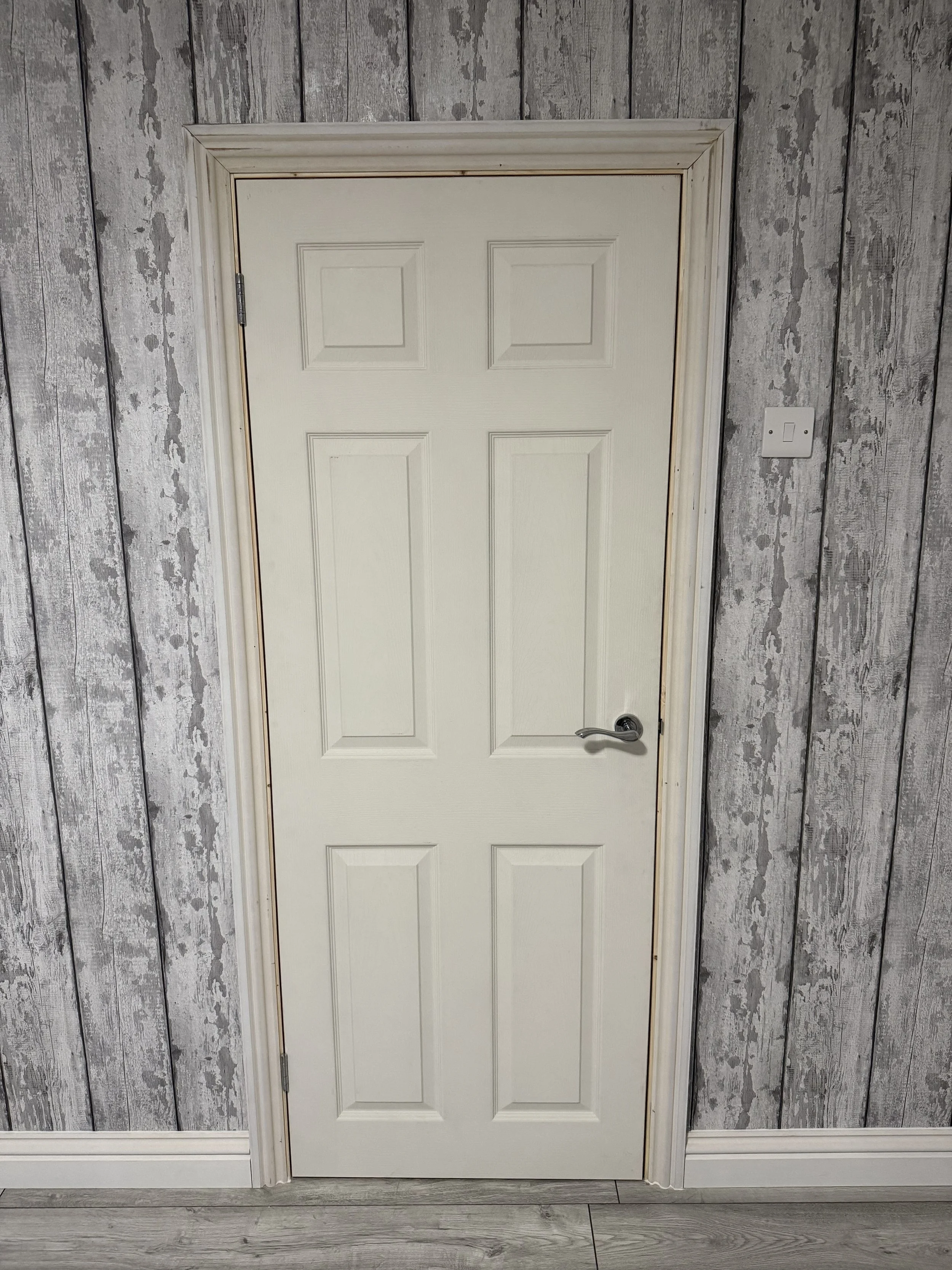 Interior view of a closed white door with a silver handle in a room with gray wooden walls and white baseboard, Wood flooring.