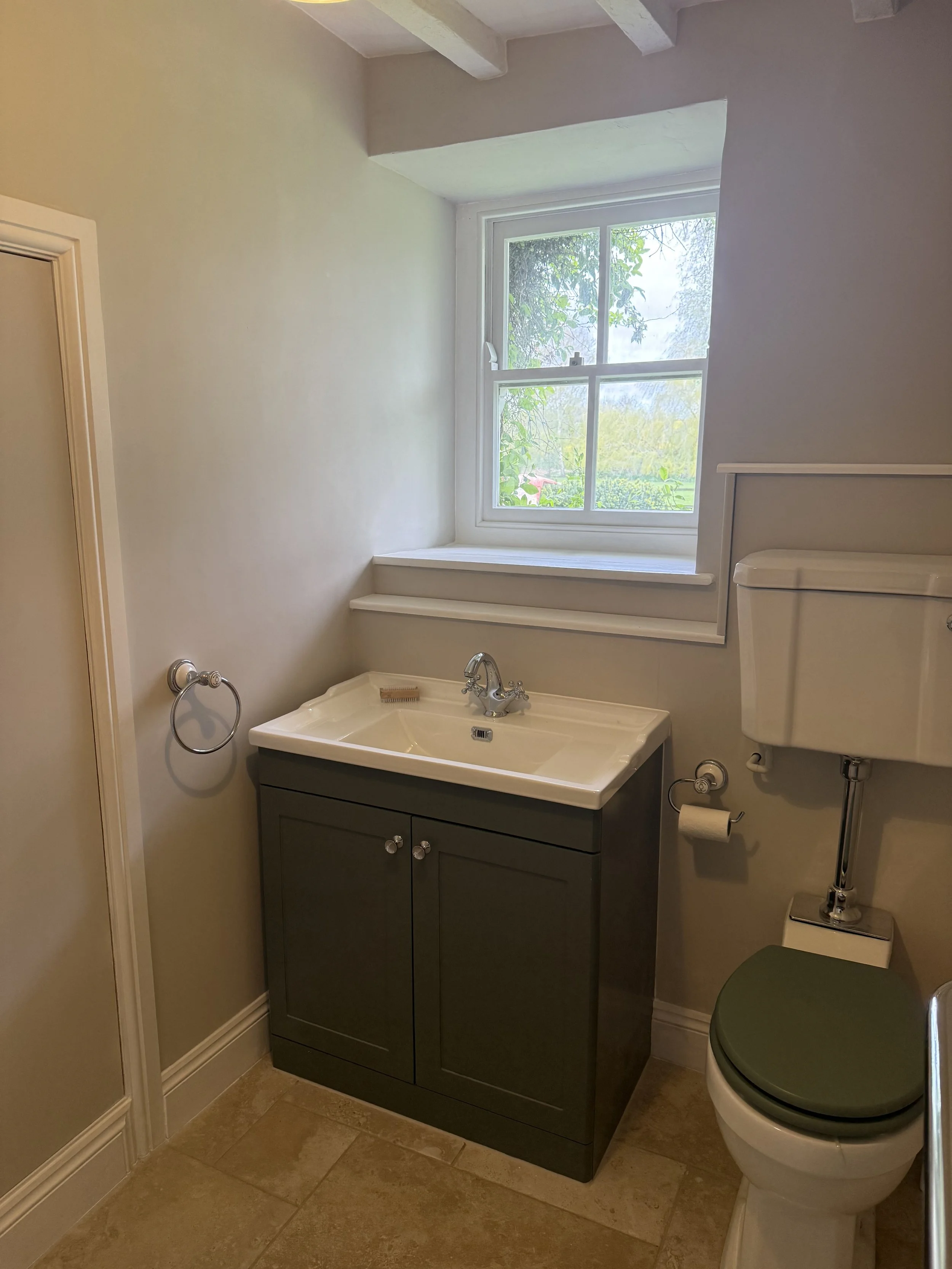 A small bathroom with a window, a sink with a dark green cabinet, a toilet with a matching green seat, and beige tiled flooring.