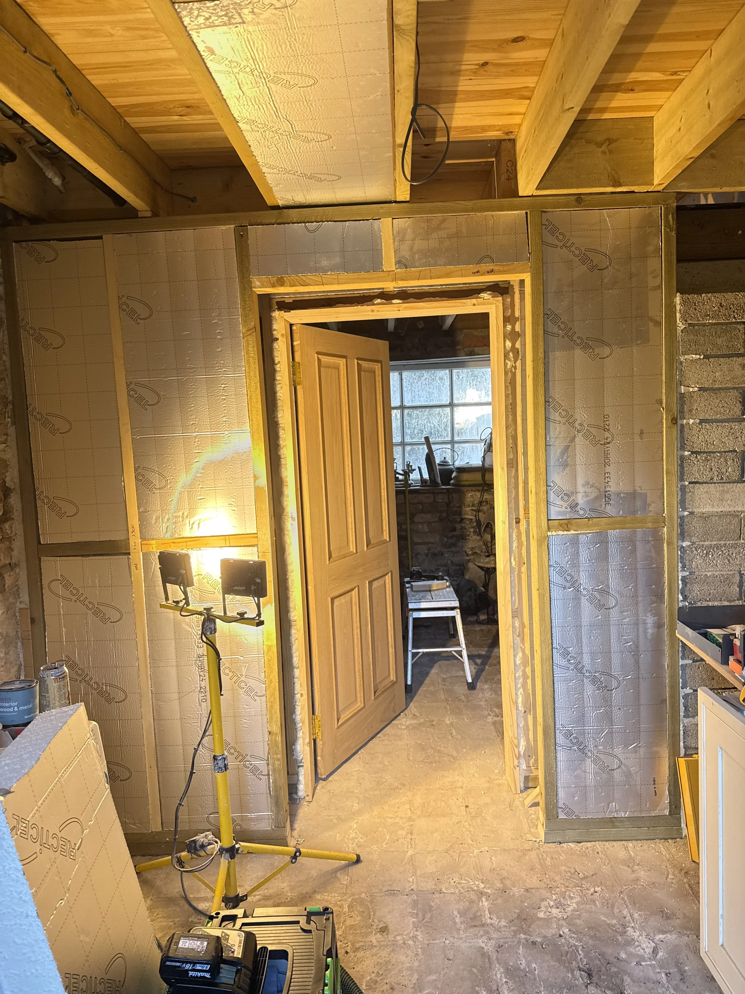 Interior view of a room under construction with exposed framing, insulation, and a partially installed door. Construction tools and lights are visible.
