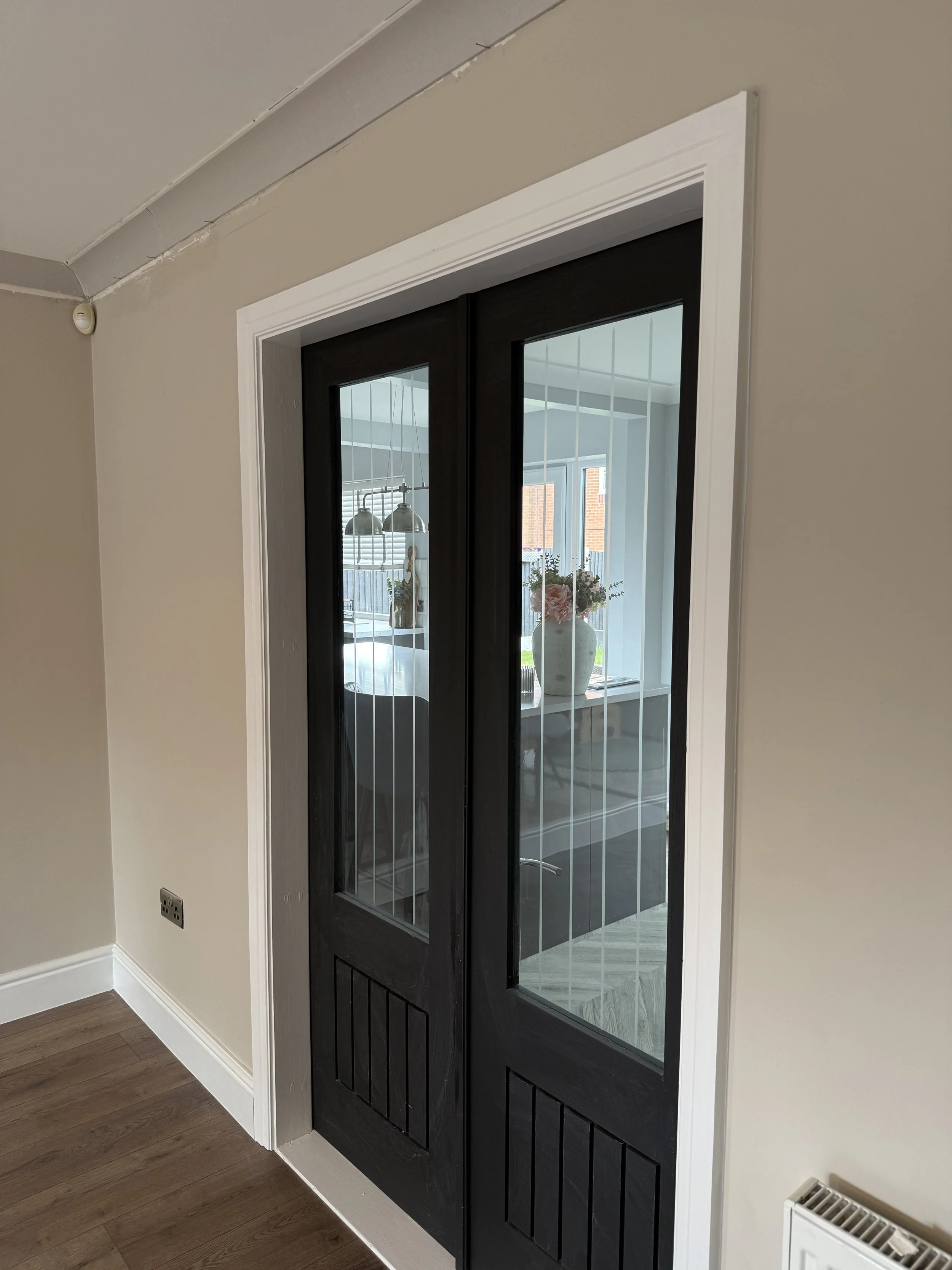 Black front door with glass panels, white trim, and wood paneling at the bottom, with a view of a vase with flowers and a window inside.