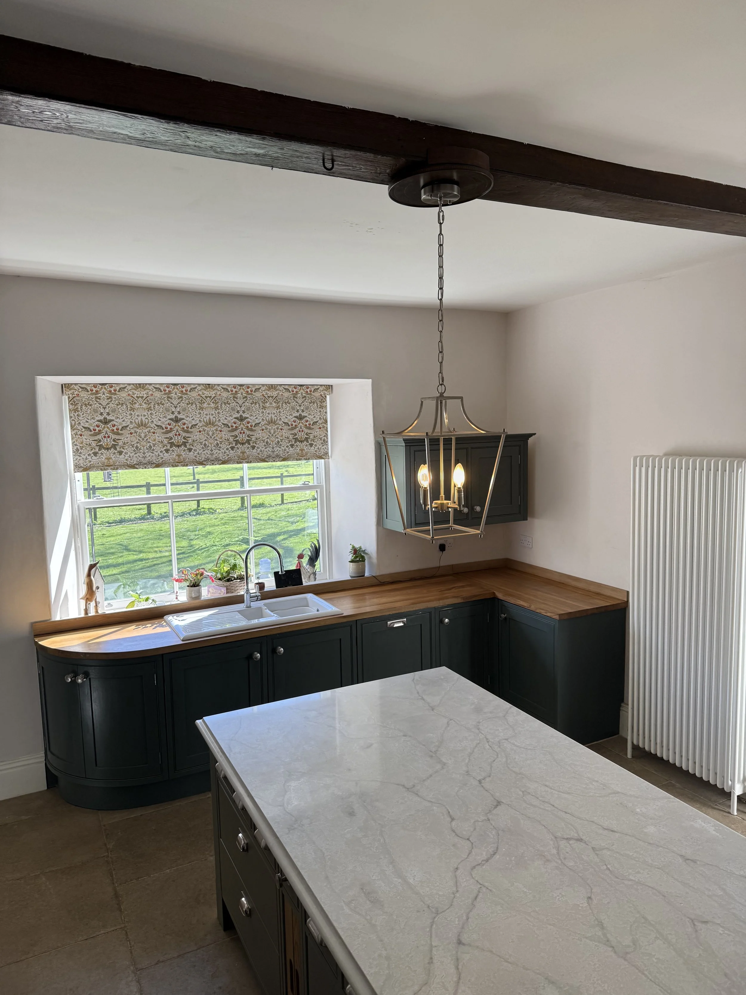 Kitchen with dark cabinetry, wooden countertops, a large window with a roman shade, a white sink, a hanging chandelier, and a white radiator.