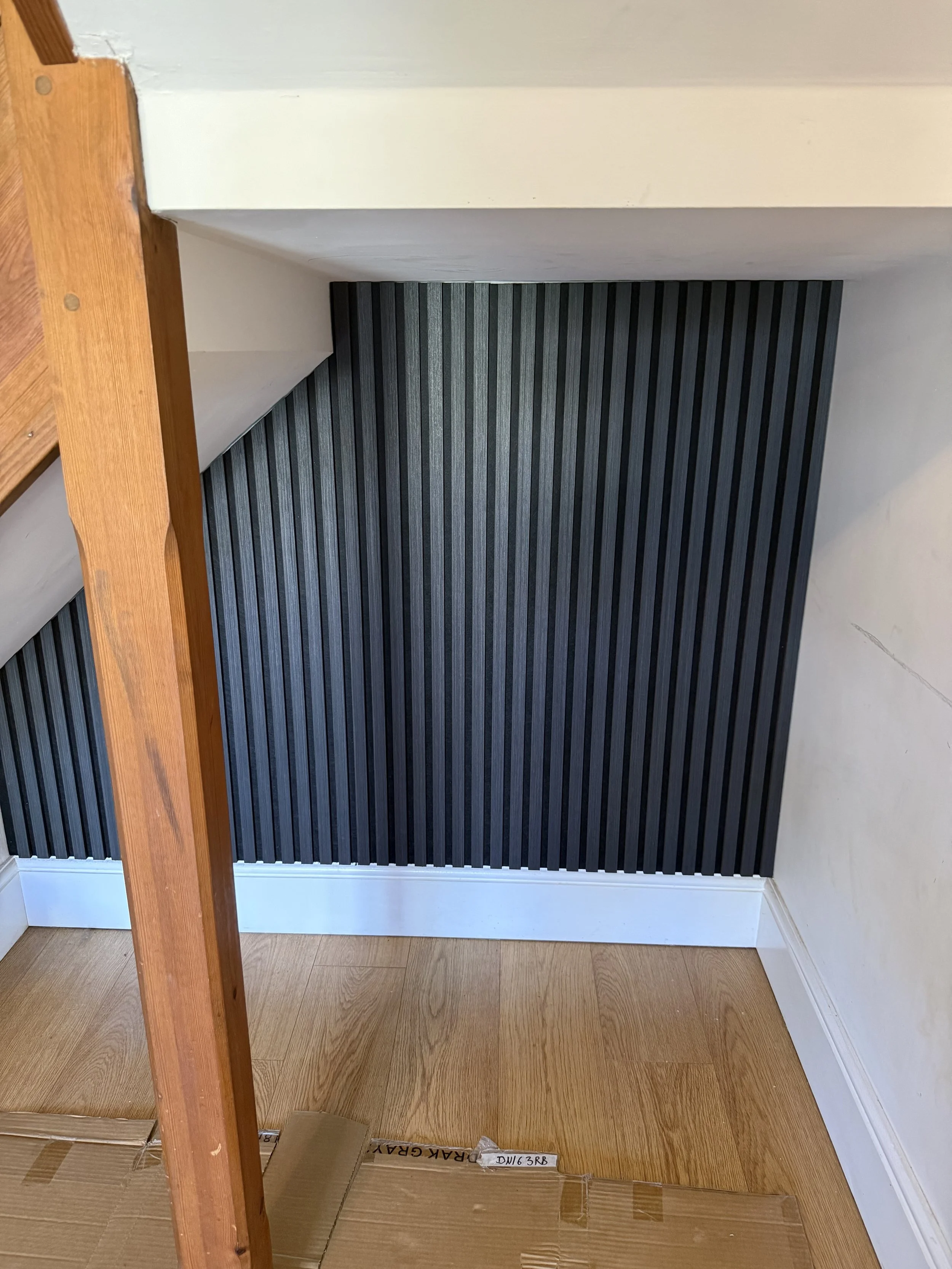 An interior corner of a room with black vertically striped wallpaper, a white baseboard, a wooden floor, and part of a wooden staircase or post.