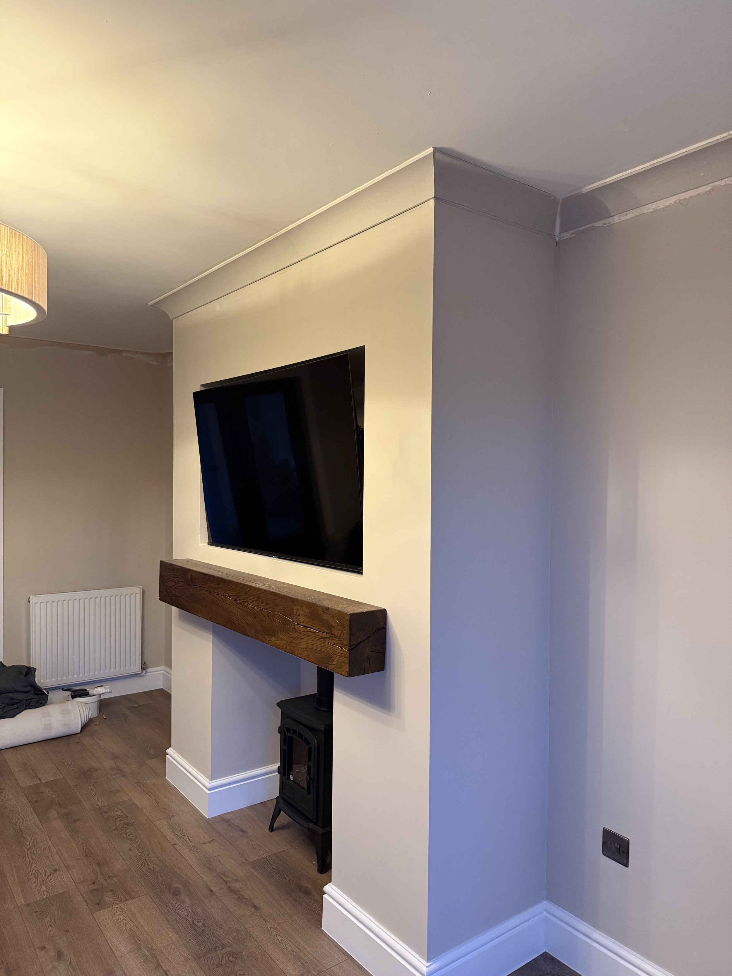 A corner of a living room with a mounted flat-screen TV on a white wall, a rustic wooden mantel, a black stove or oven below, a radiator in the corner, and hardwood flooring.