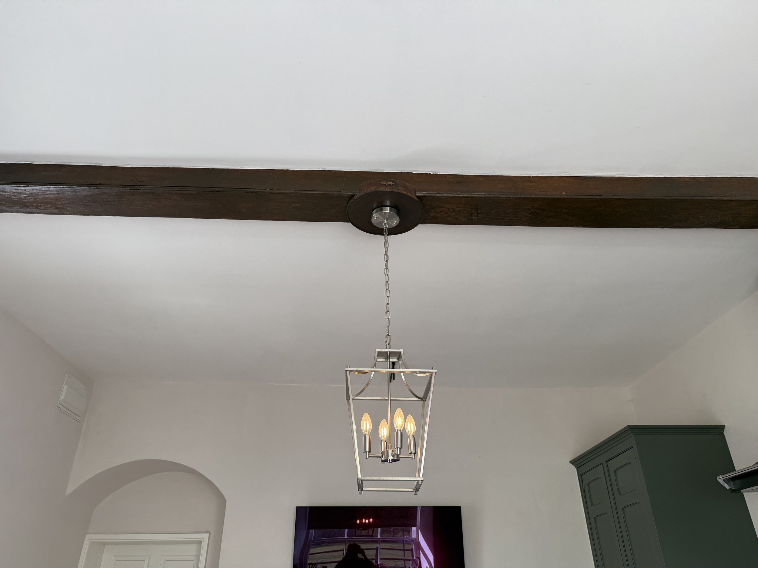 A modern chandelier with five candle-shaped light bulbs hanging from a chain attached to a wooden ceiling beam in a room with white walls and a green cabinet.
