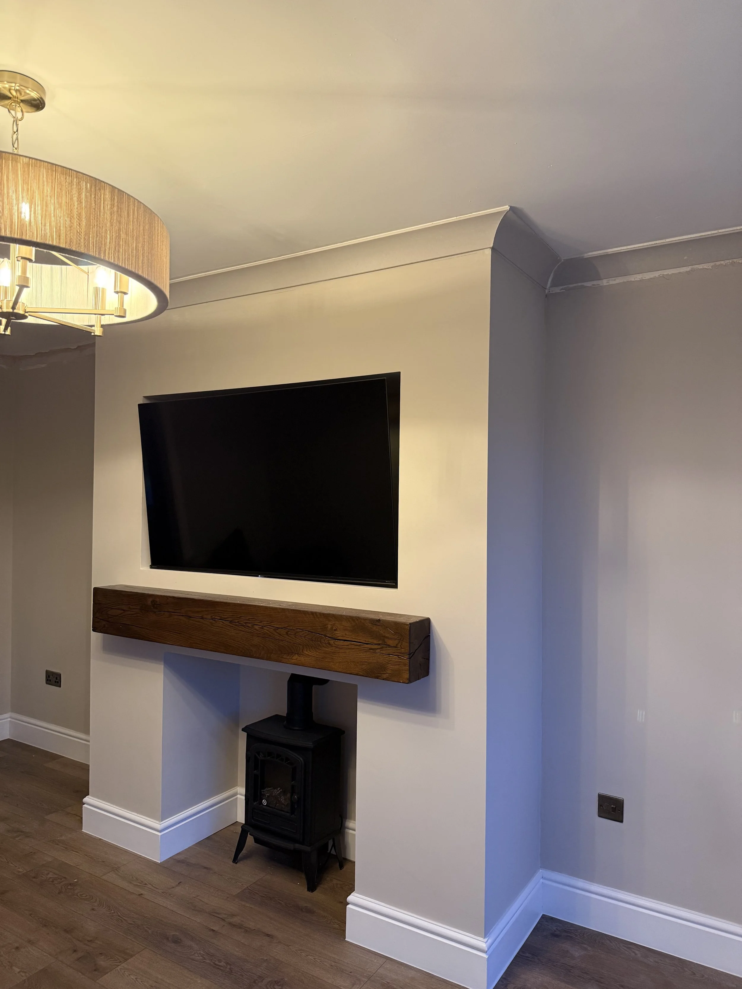 A living room corner with a wall-mounted flat-screen TV, a wooden floating shelf below, a small black stove or heater beneath the shelf, a wall mounted light fixture on the ceiling, and a corner of a blue-painted wall with crown molding.