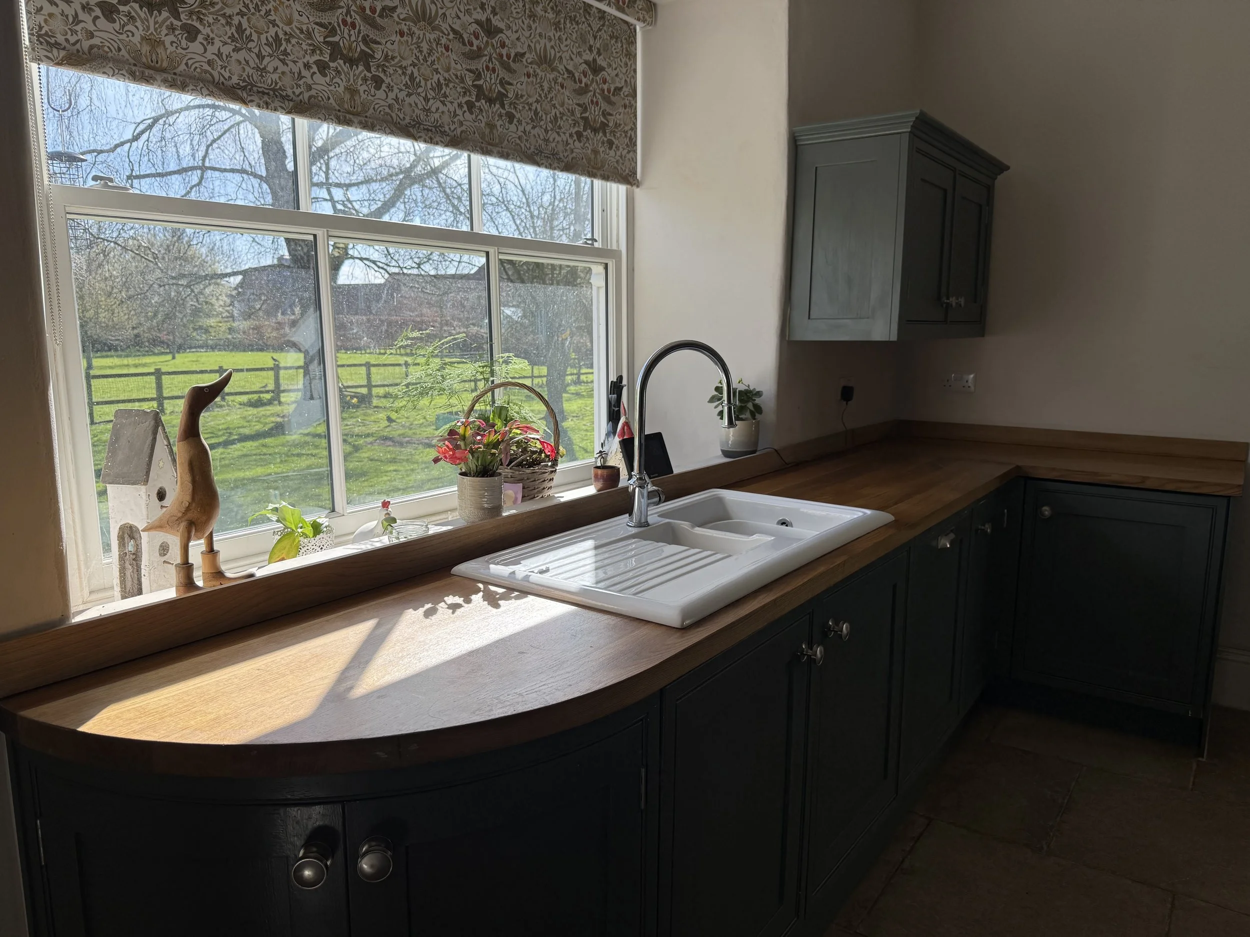 Kitchen sink with a window overlooking a green yard with trees and a fence, decorated with potted plants and a wooden bird figurine.