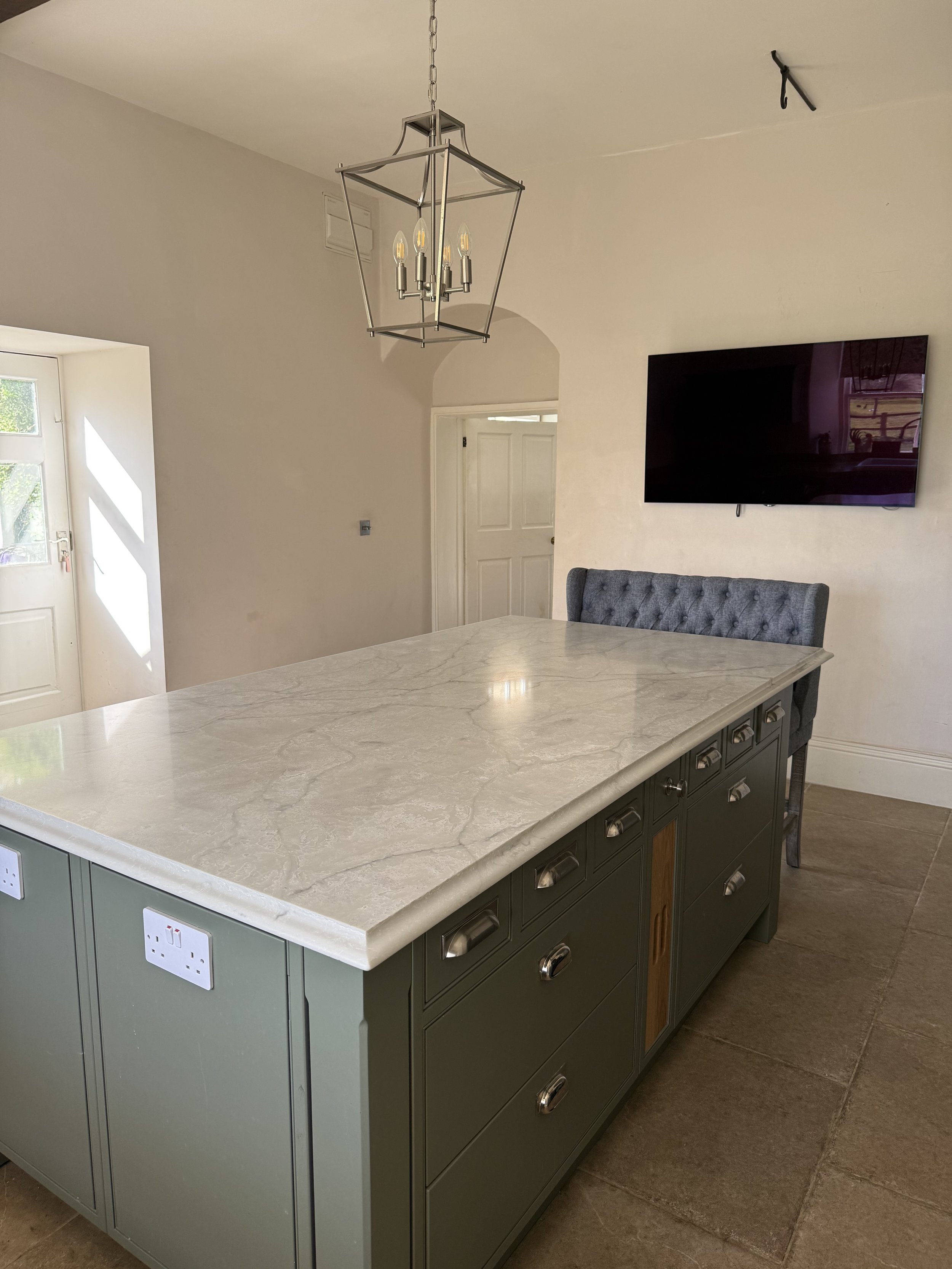 A modern kitchen with a green island featuring a marble countertop, electrical outlets, and multiple drawers. There's a gray upholstered chair at the end of the island. A wall-mounted flat-screen TV is on the wall, and a hanging geometric lantern-sty