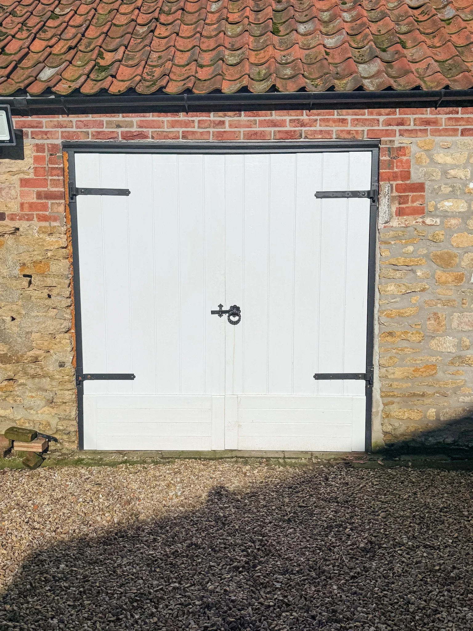 A white wooden garage door with black hinges and latch, set in a brick and stone wall. The roof above has red tiles, and the ground in front is gravel.