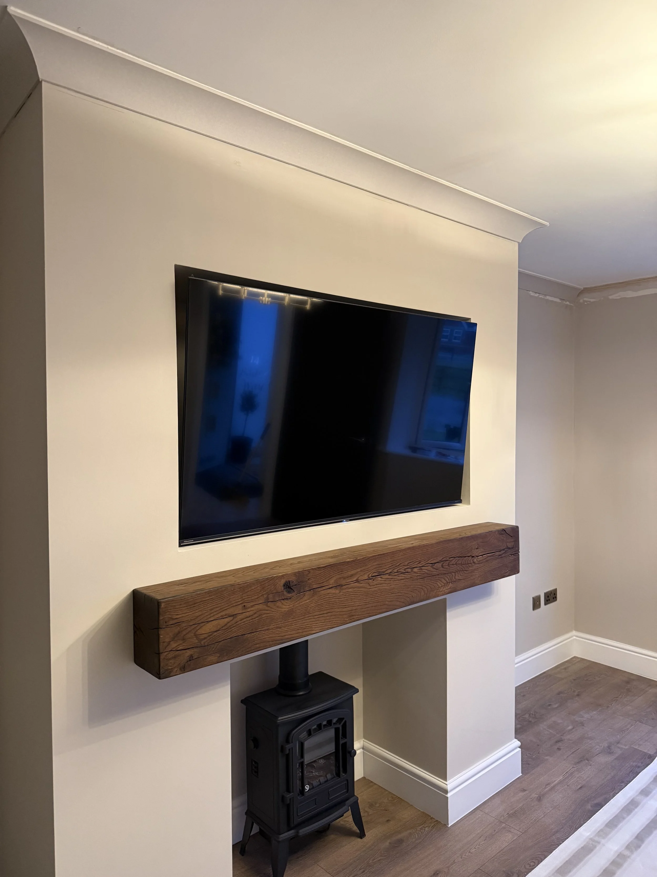 Living room wall with mounted flat-screen TV, wooden shelf, and decorative stove on light hardwood floor.