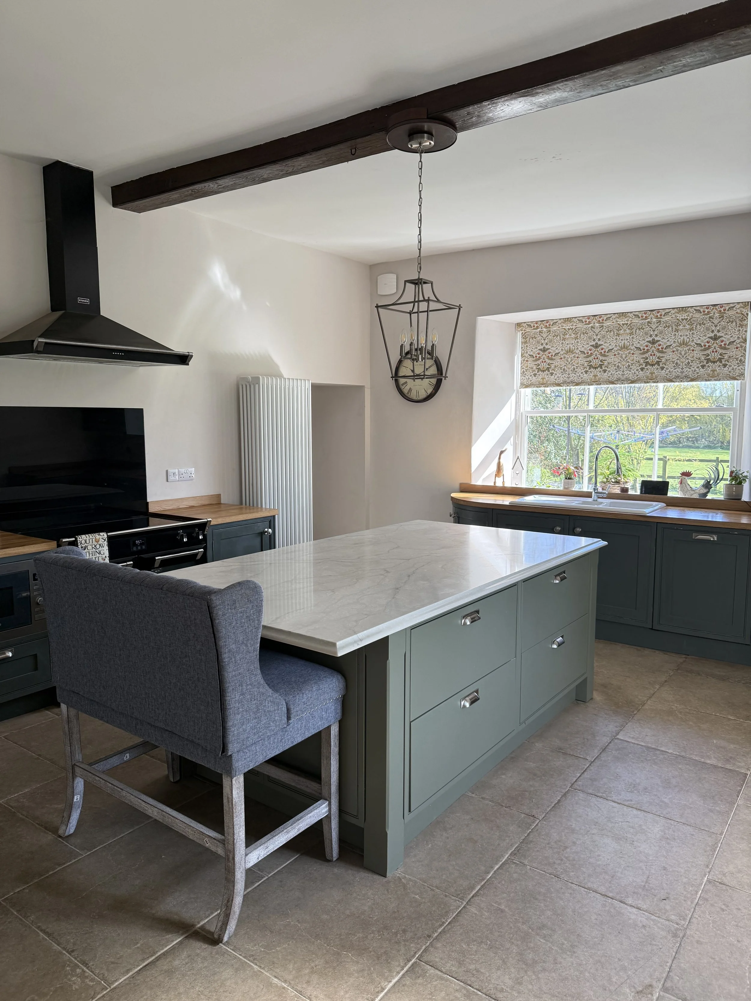 Modern kitchen with a green kitchen island, gray upholstered chair, large window with floral curtain, black oven range, wooden countertops, and a black extractor hood. Decorative clock with chandelier-style light fixture hanging overhead.