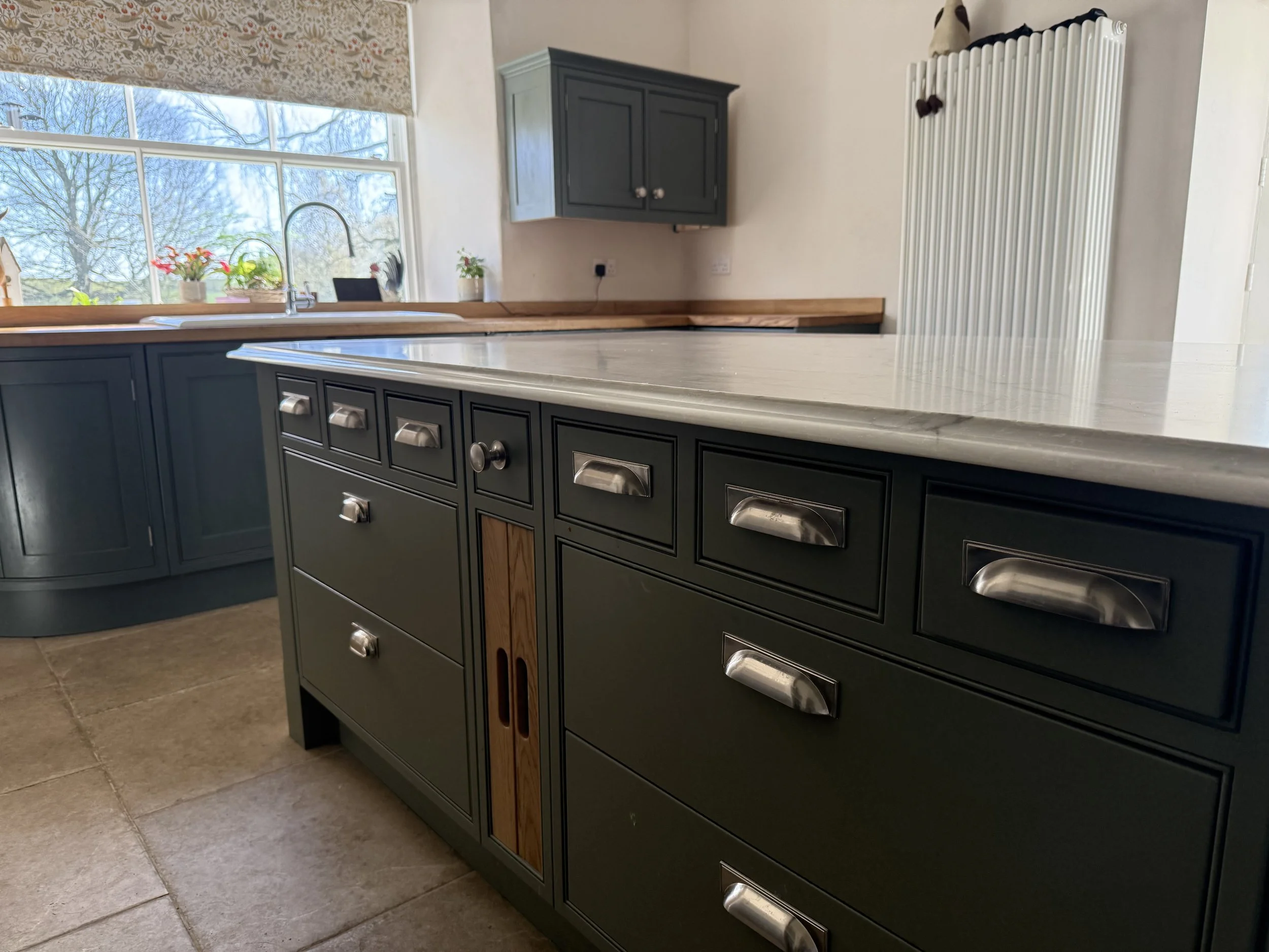A kitchen with green cabinetry, a marble countertop, and a window above the sink showing trees outside. There are small plants on the windowsill, and a radiator on the wall to the right.