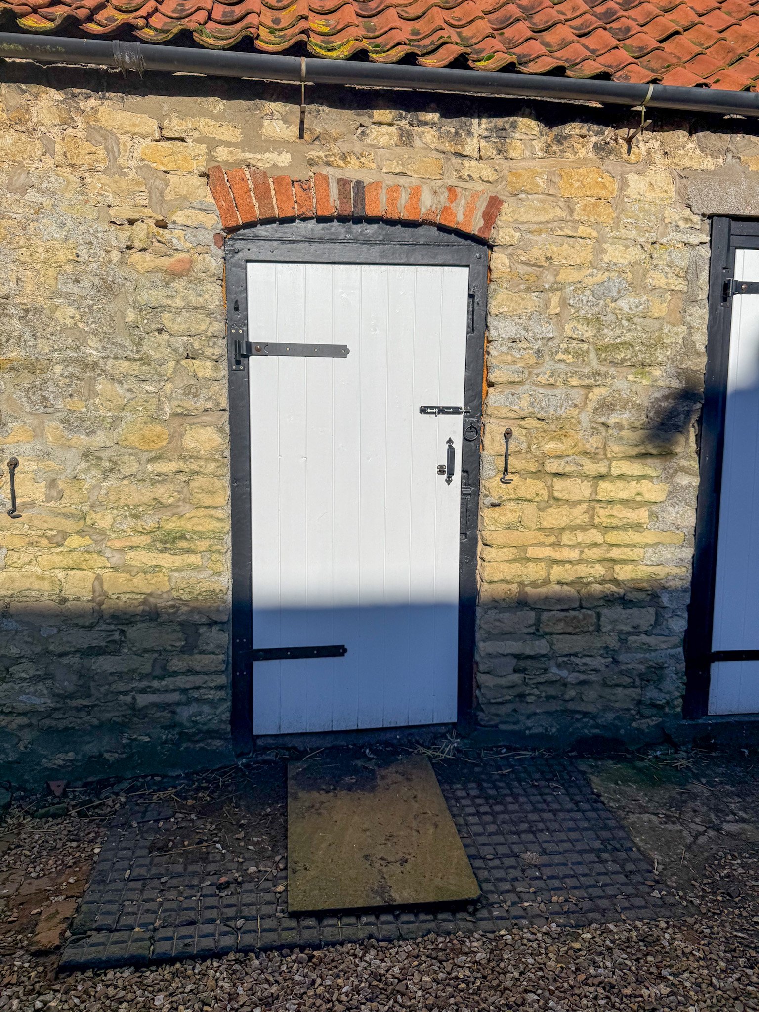 A small white door with black hinges and a latch in a rustic stone wall, with a wooden board on the ground in front of it. The wall has a brick arch above the door, and a shadow partially covers the lower part of the door.