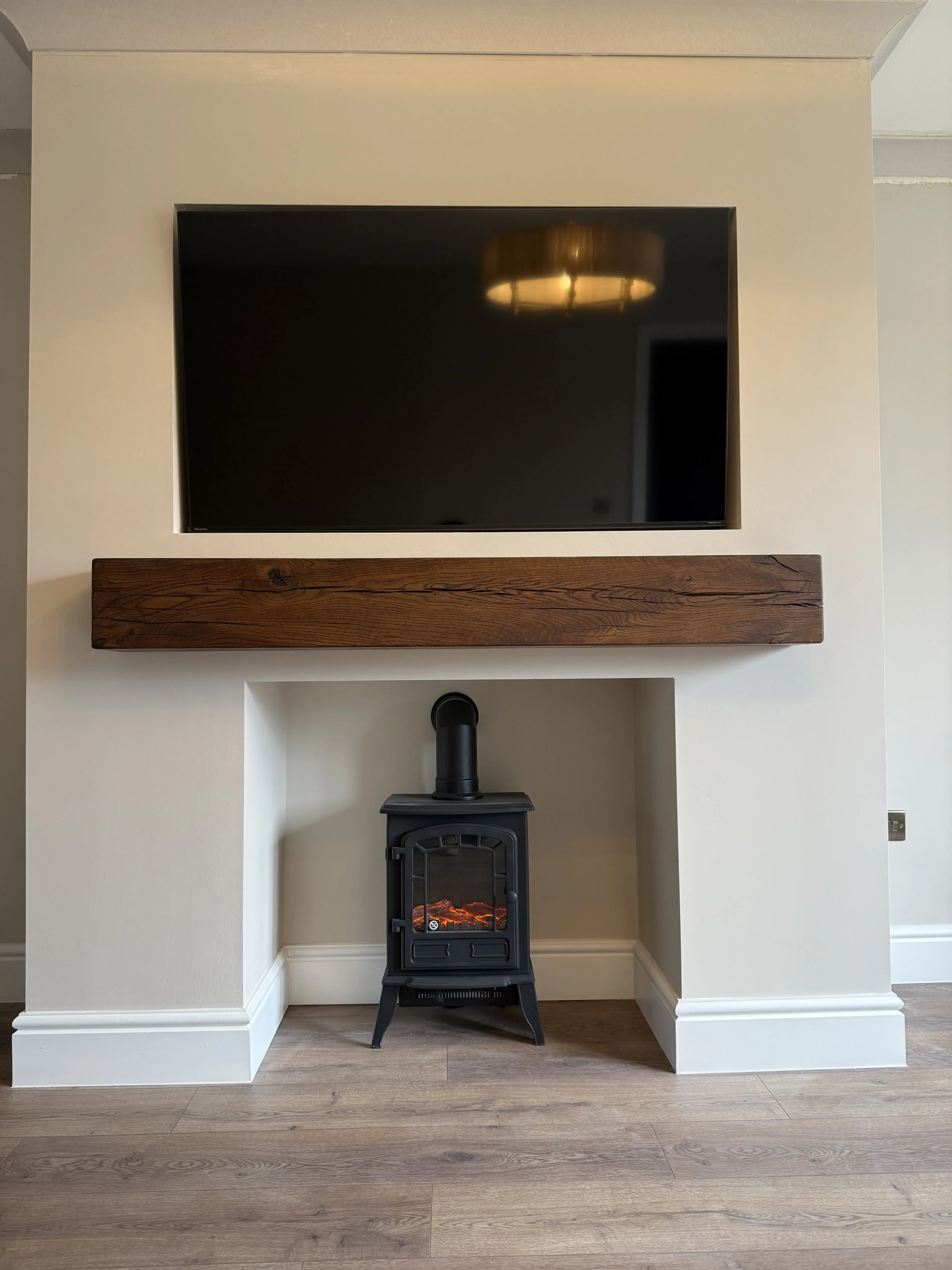 A room with a mounted flat-screen TV on the wall, above a wooden mantel, and a black wood stove with a visible fire inside below the mantel, all set against a beige wall and wooden floor.