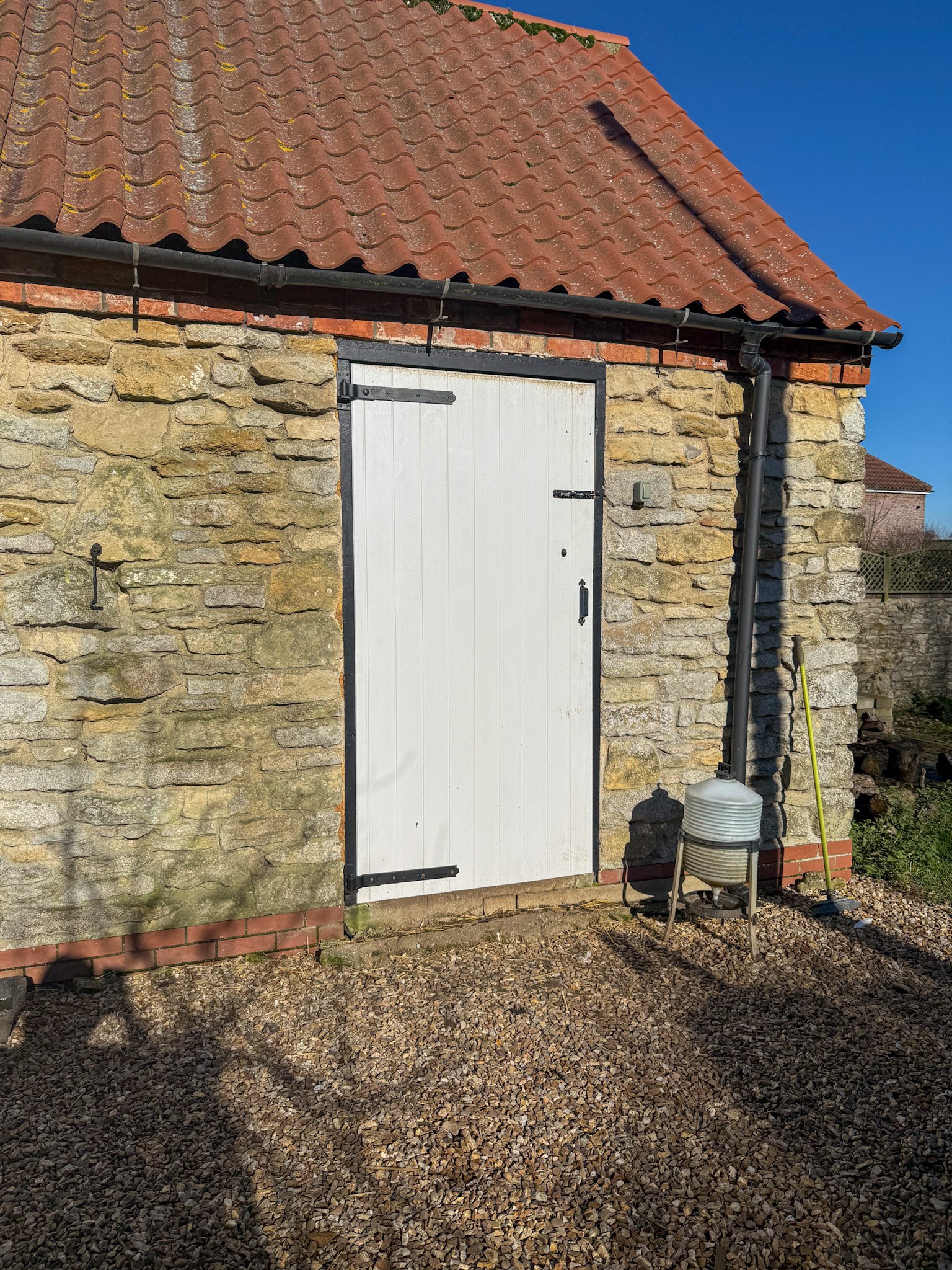 Stone building with a white door, red tiled roof, and black gutter. Gravel ground with shadows, nearby gardening tools, and a blue sky.