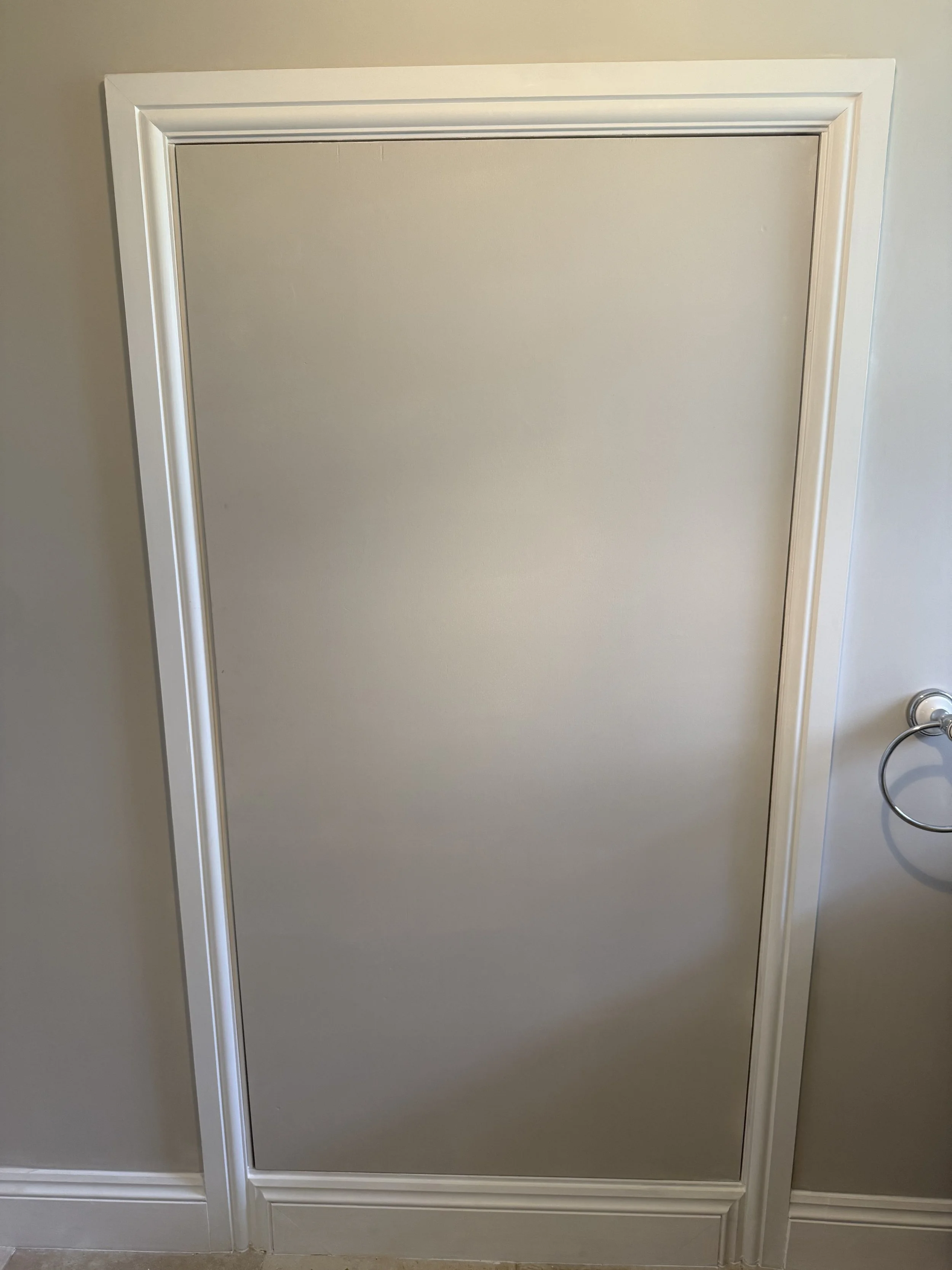 A closed beige door with white trim in a room with light-colored walls and a towel ring attached to the wall on the right side.