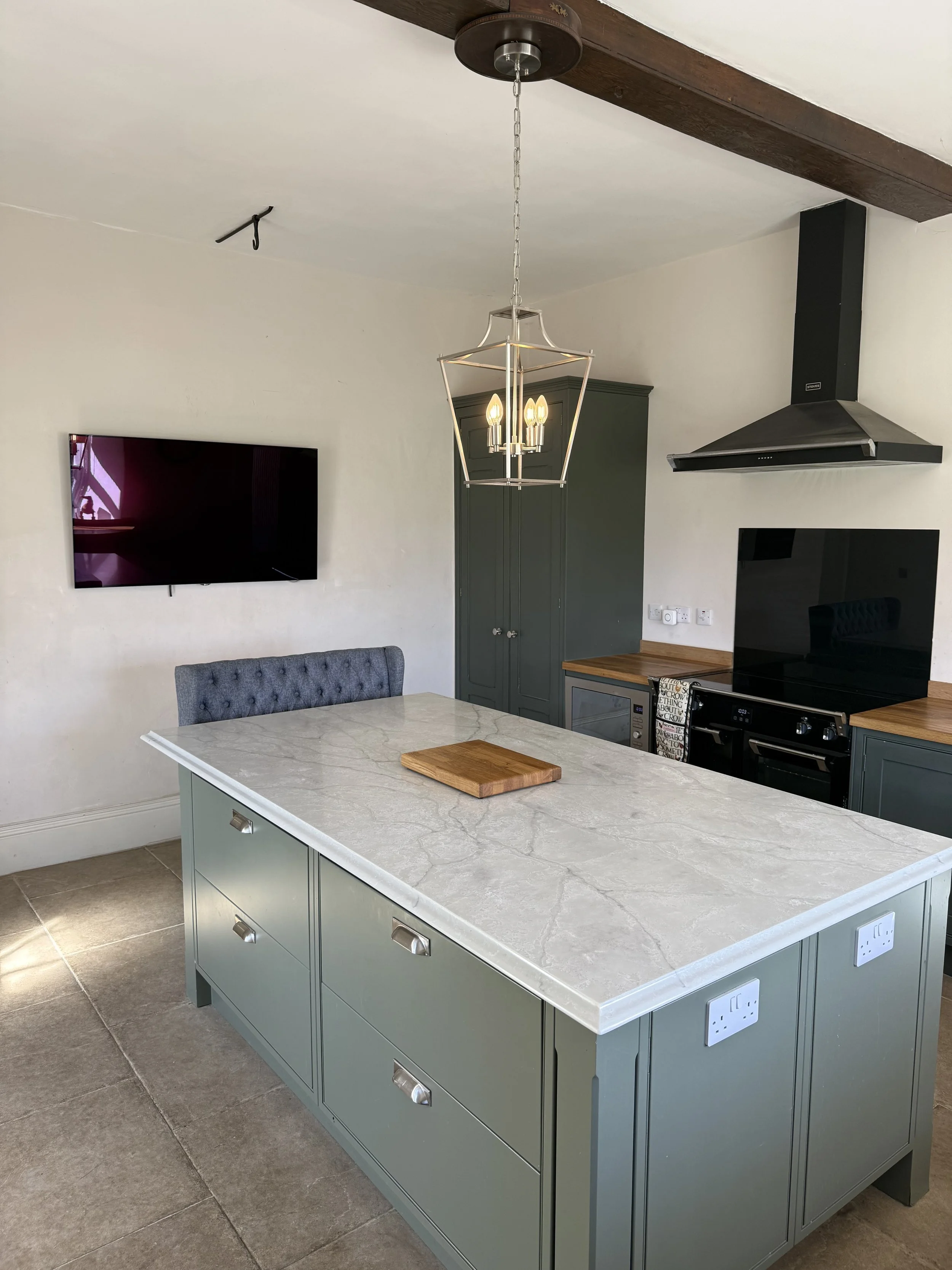 Modern kitchen with green cabinets, a central island with a marble countertop, a hanging chandelier, a wall-mounted TV, and a built-in oven with a black range hood.