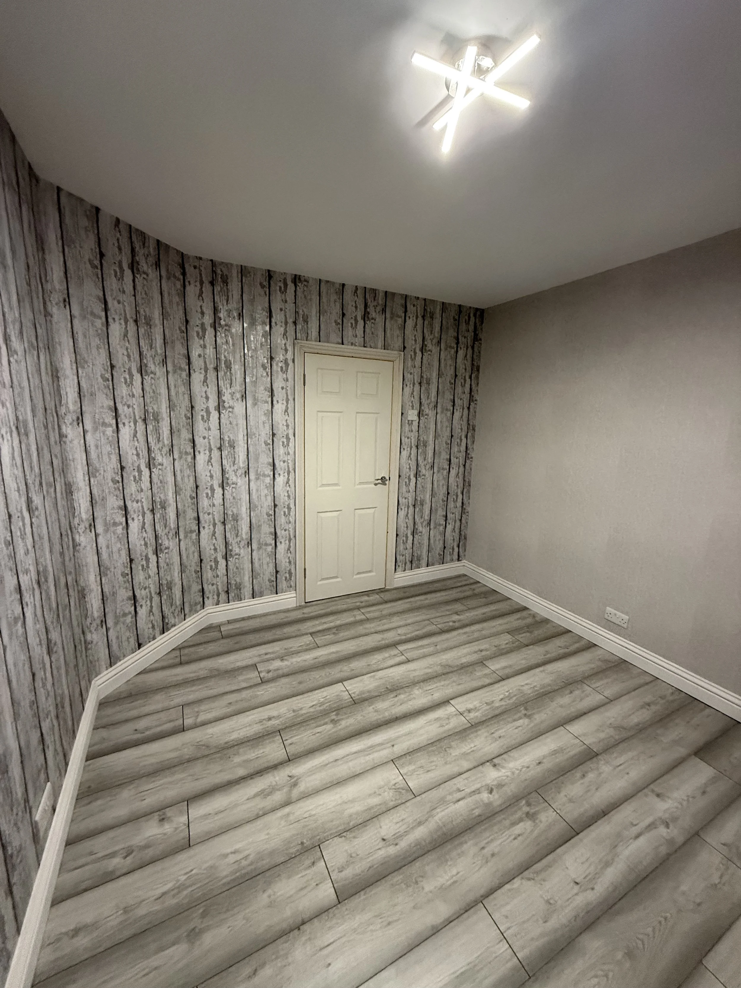 Interior of a room with wood-paneled accent wall, light-colored flooring, a small white door, and a modern ceiling light fixture.