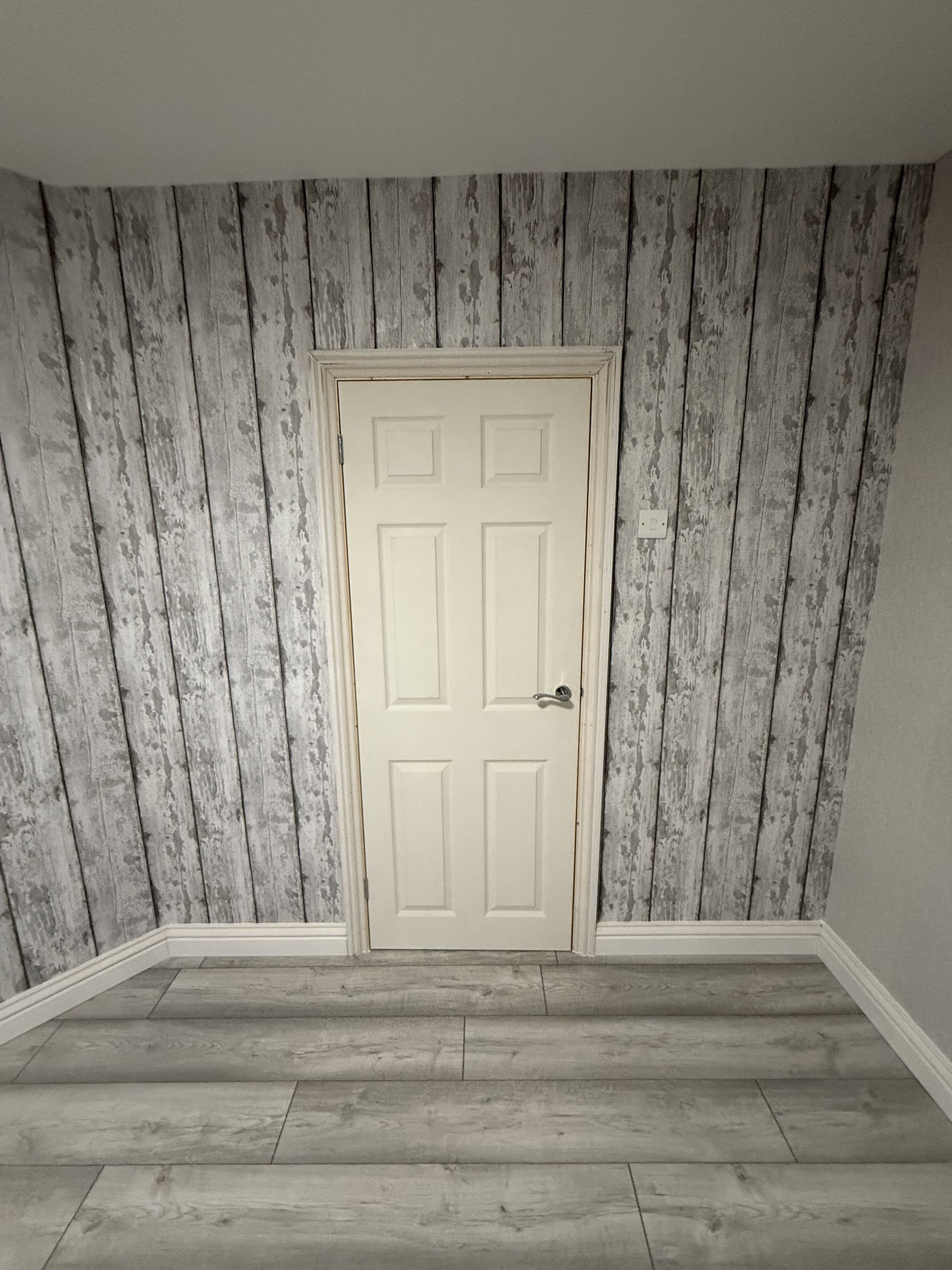 A door set in a wood-paneled wall with gray and white tones, located in a room with light-colored wood flooring and white baseboards.