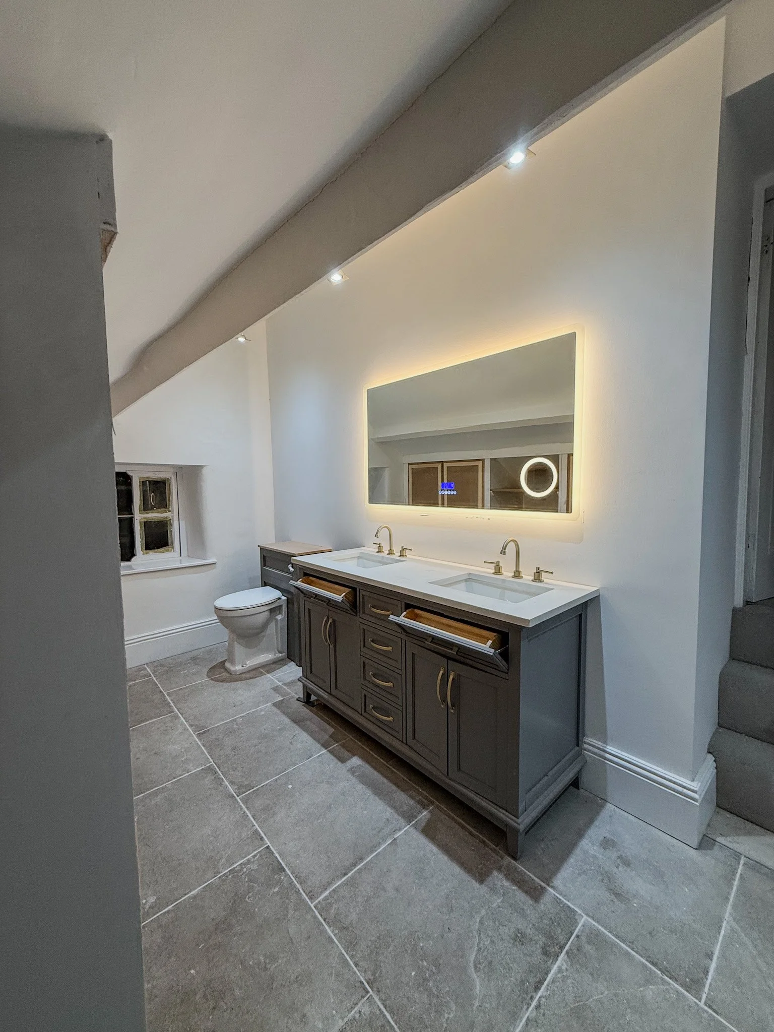 Bathroom with double vanity sink, illuminated mirror, toilet, small window, and gray tiled floor.