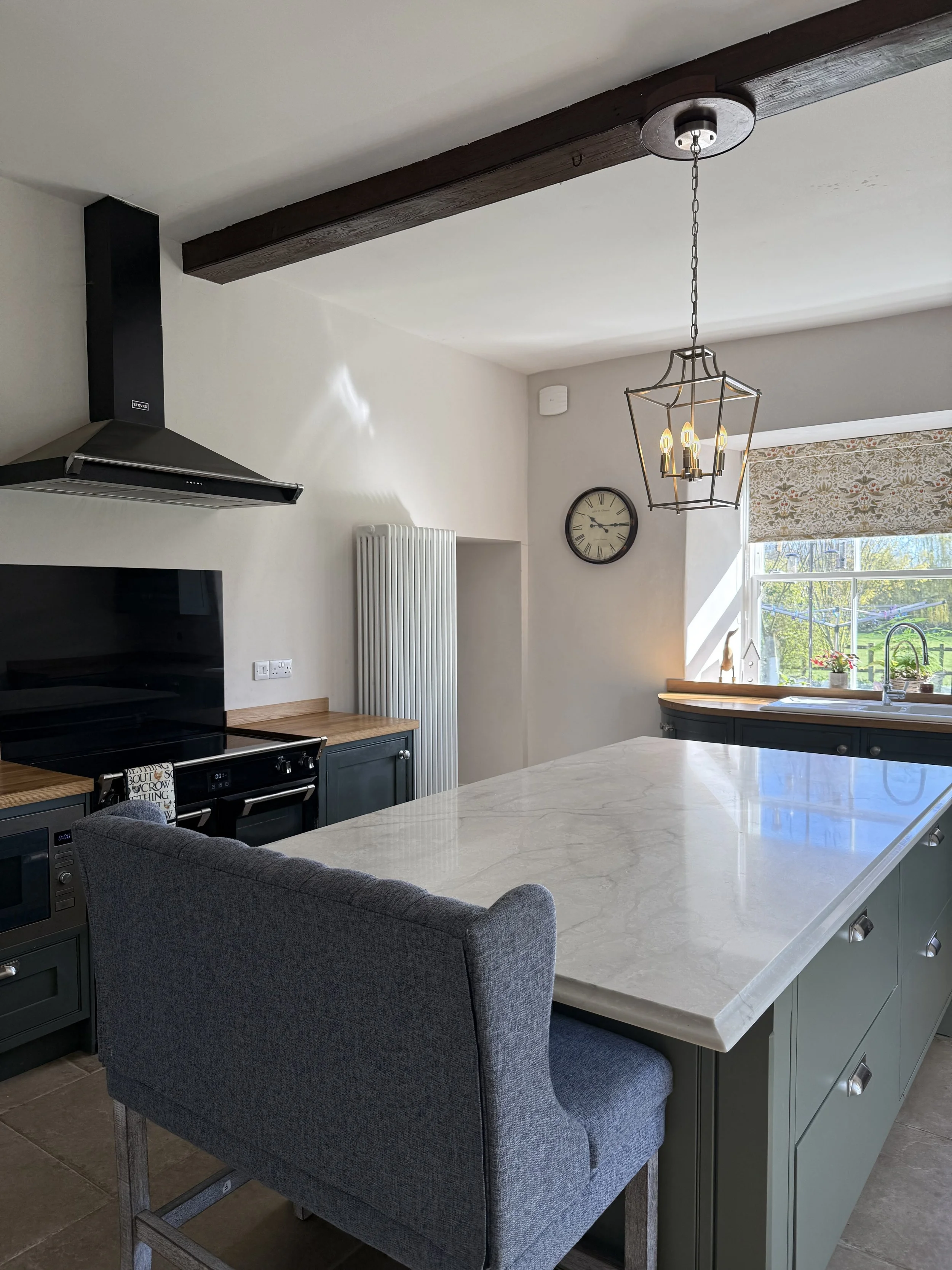 Modern kitchen with gray cabinets, a large white marble island, a wall clock, a window with a floral valance, and a chandelier lighting fixture.