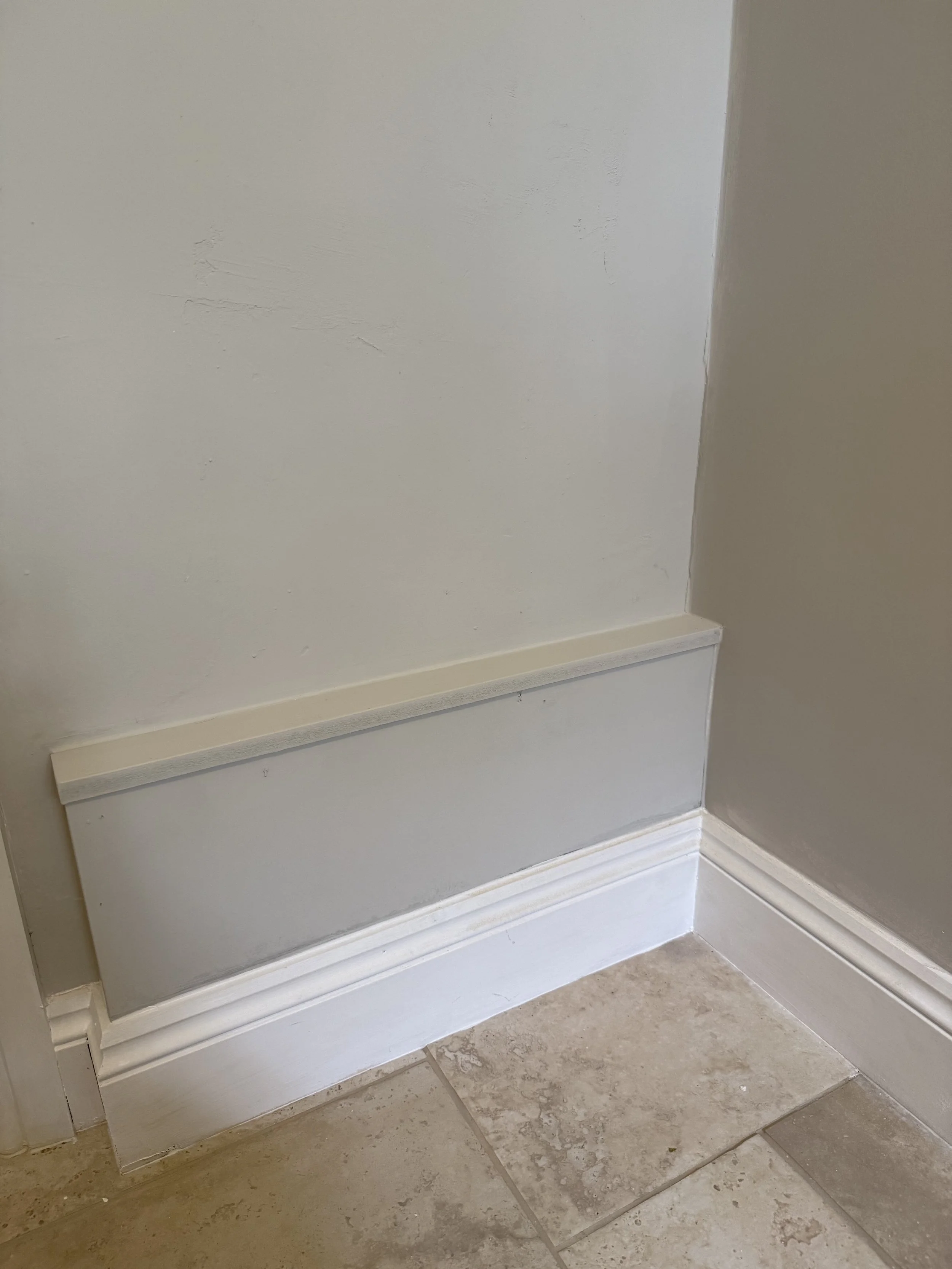 Interior corner of a wall with a white trim or baseboard, tiled floor, and wall sconces painted in light and darker shades of beige.