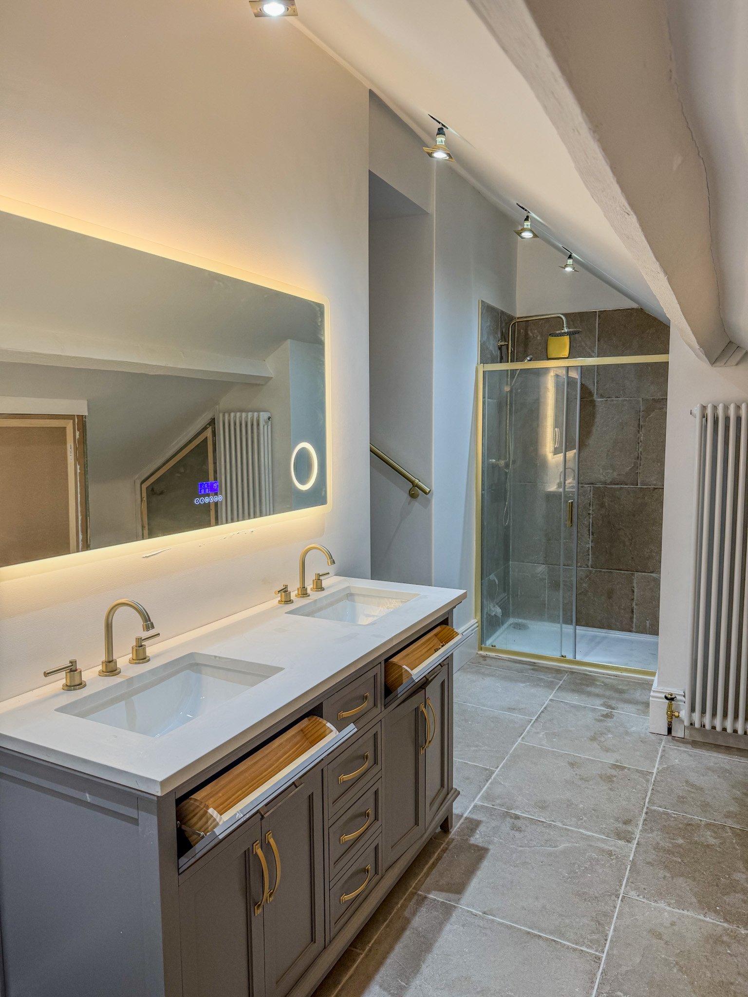 Modern bathroom with double sink vanity, large backlit mirror, walk-in shower with glass door, beige and gray tiles, and white walls.