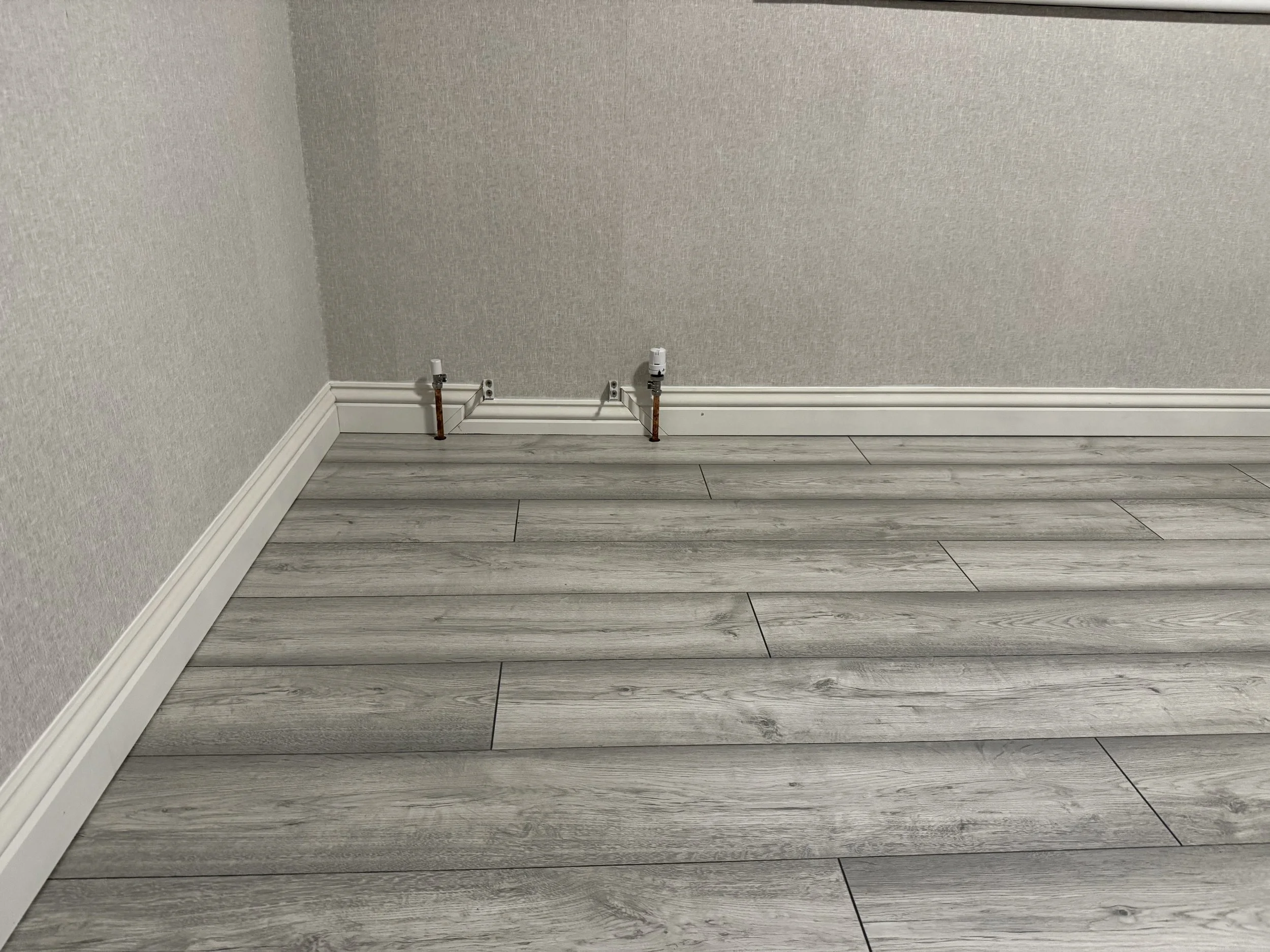 Empty corner of a room with two exposed water pipes coming out of the wall, a white baseboard, and light-colored wood laminate flooring.