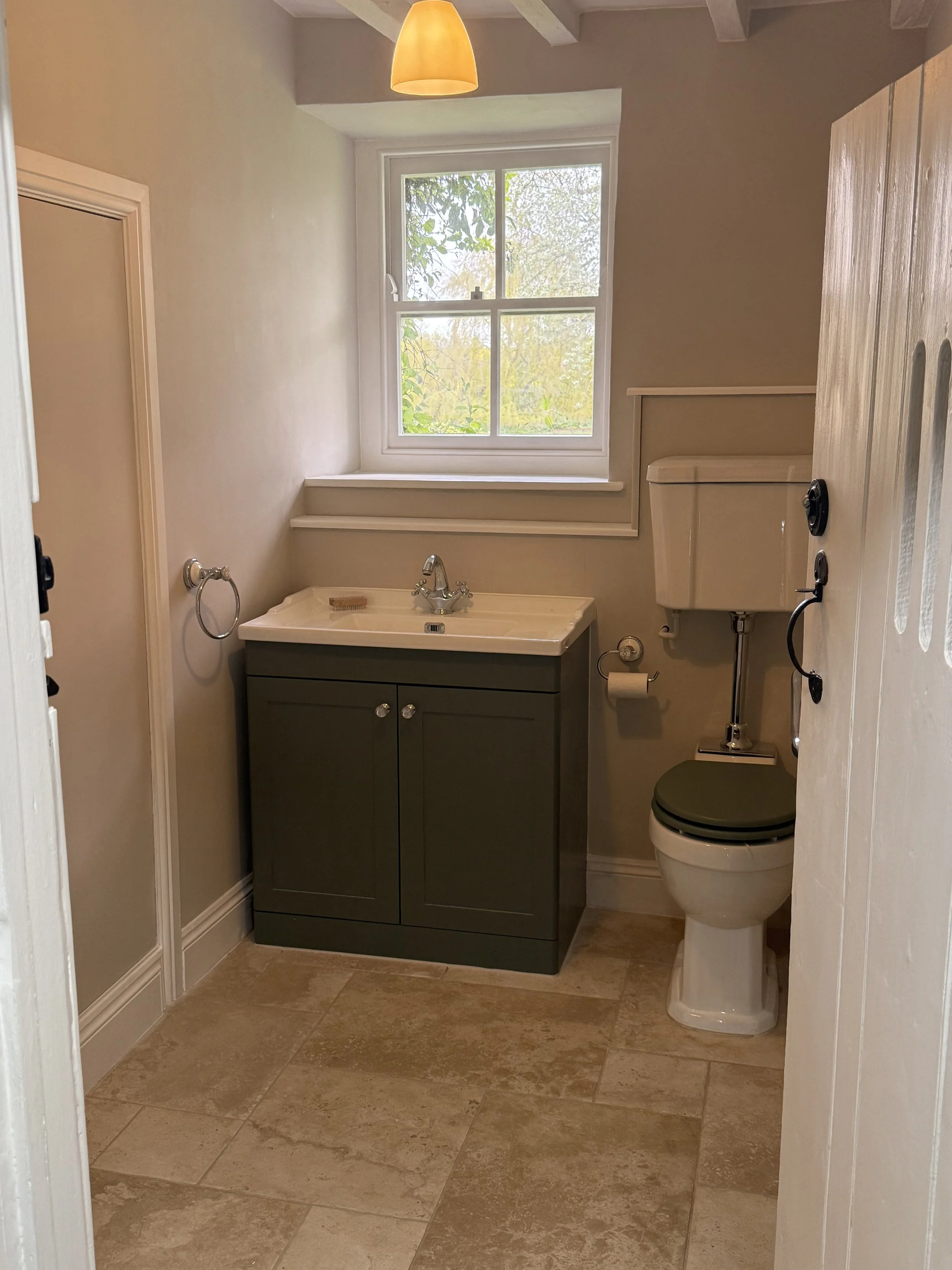 A small bathroom with a window, beige tiled floor, green vanity with a sink, and a toilet with a green seat. There is a yellow lampshade hanging from the ceiling.