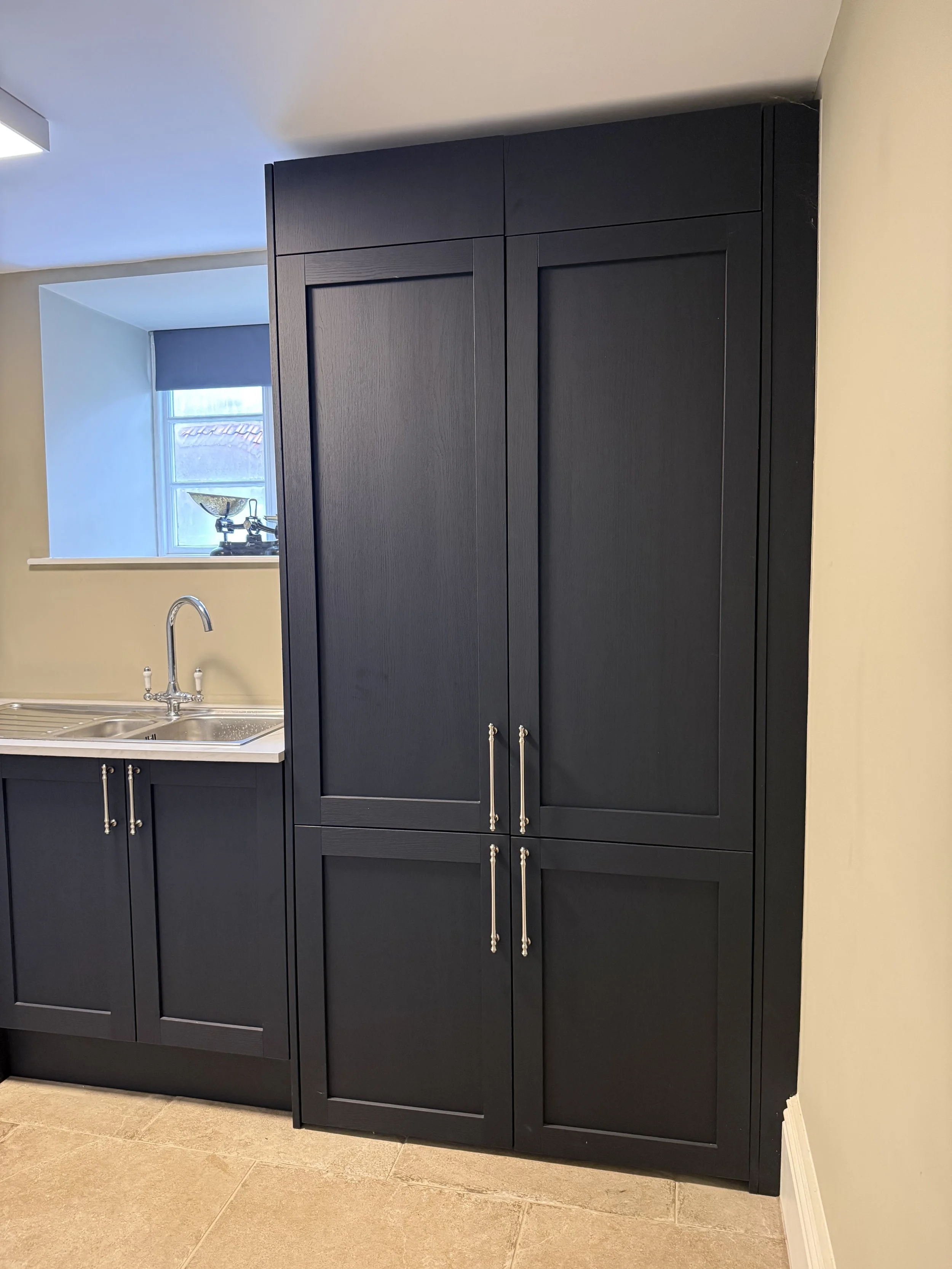 Dark wooden kitchen cabinet with silver handles in a room with beige tiled floor and a window with blinds and a decorative bowl on the sill.