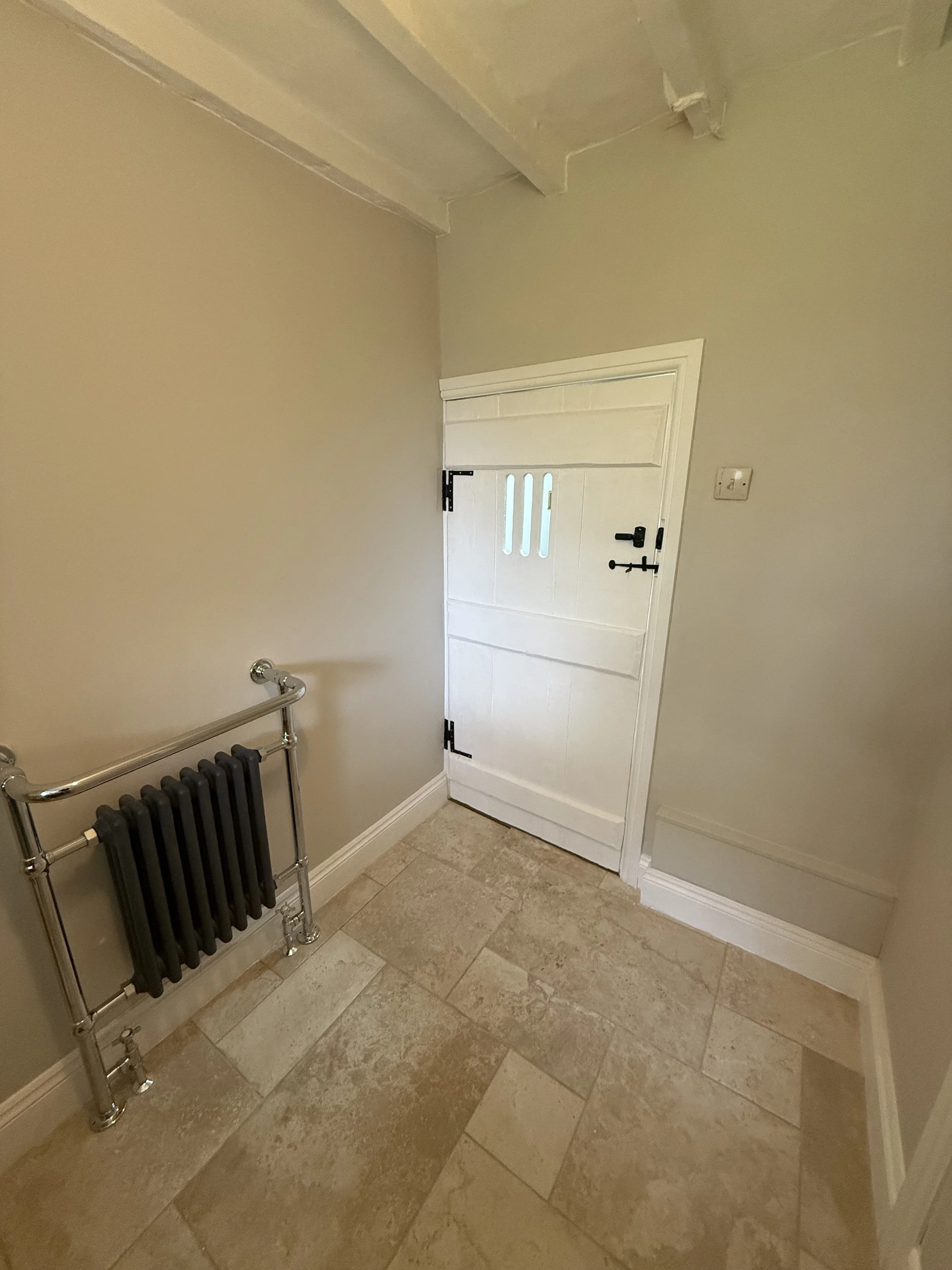Small room with beige tiled floor, a white door with three rectangular windows, a black latch, and a black lock, and a chrome radiator on the left wall.