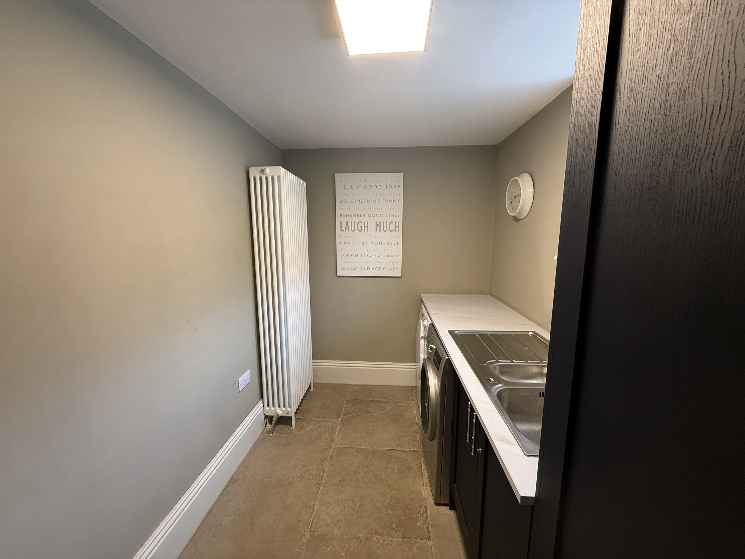 Small laundry room with a grey wall, a white radiator, a poster encouraging laughter, a white countertop with a stainless steel sink, and a compact washing machine.