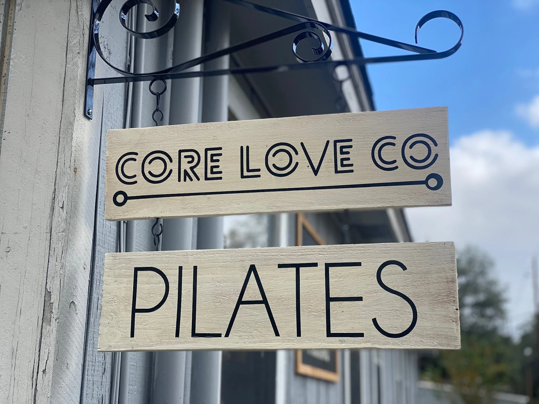 Wooden sign with black painted text that reads 'CORE LOVE CO' and part of the word 'PLACES' visible underneath. The sign is hanging from a black metal hook attached to a weathered white wooden surface, with sky and some building elements in the background.