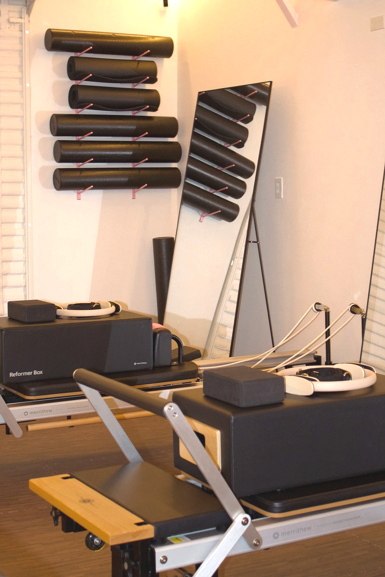 Fitness equipment including black foam rollers mounted on a wall, a mirror, and a wooden Pilates reformer machine with black padding and adjustable springs, in a well-lit exercise room.