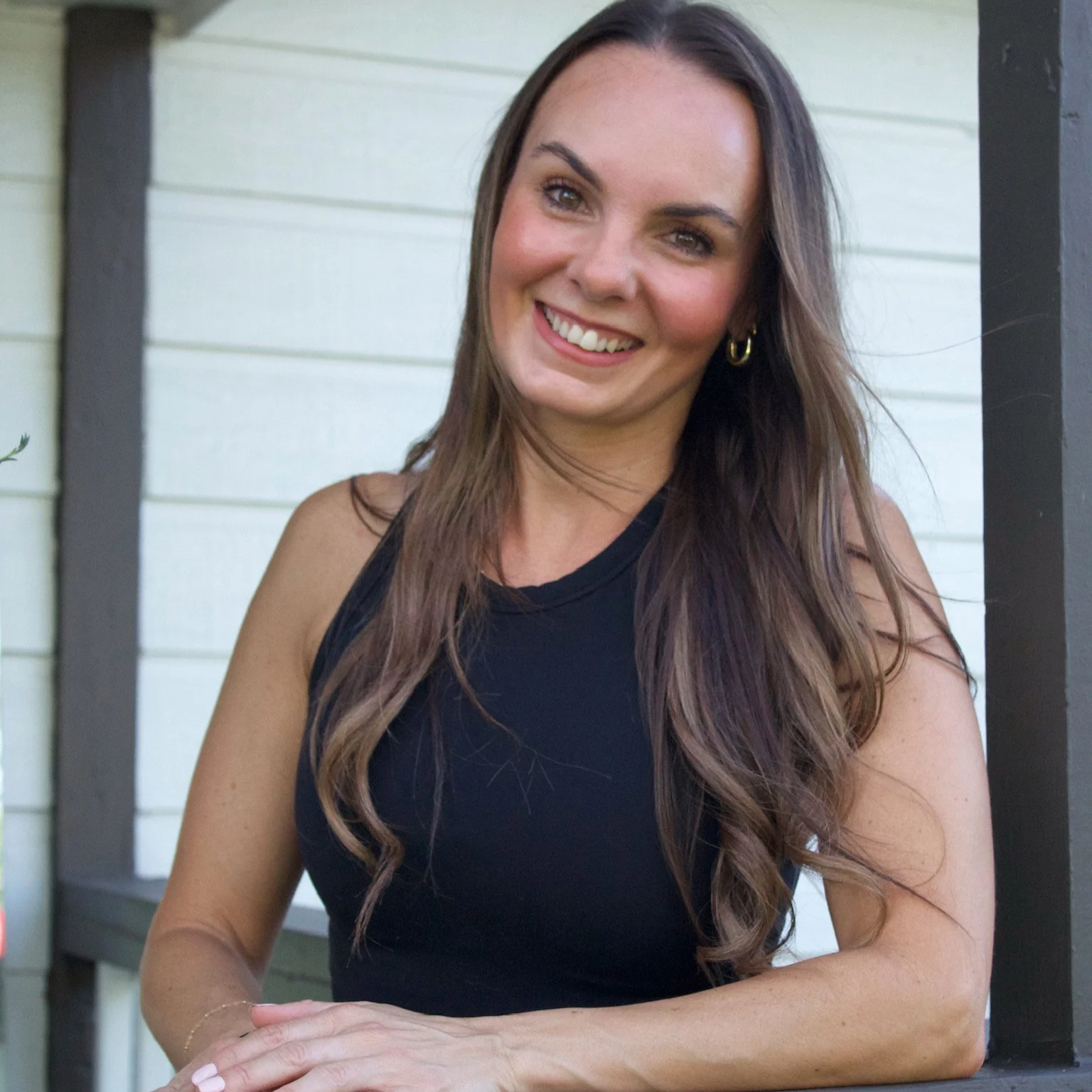 Smiling woman with long wavy brown hair wearing a black sleeveless top, sitting outdoors with a white wooden wall and dark trim in the background.