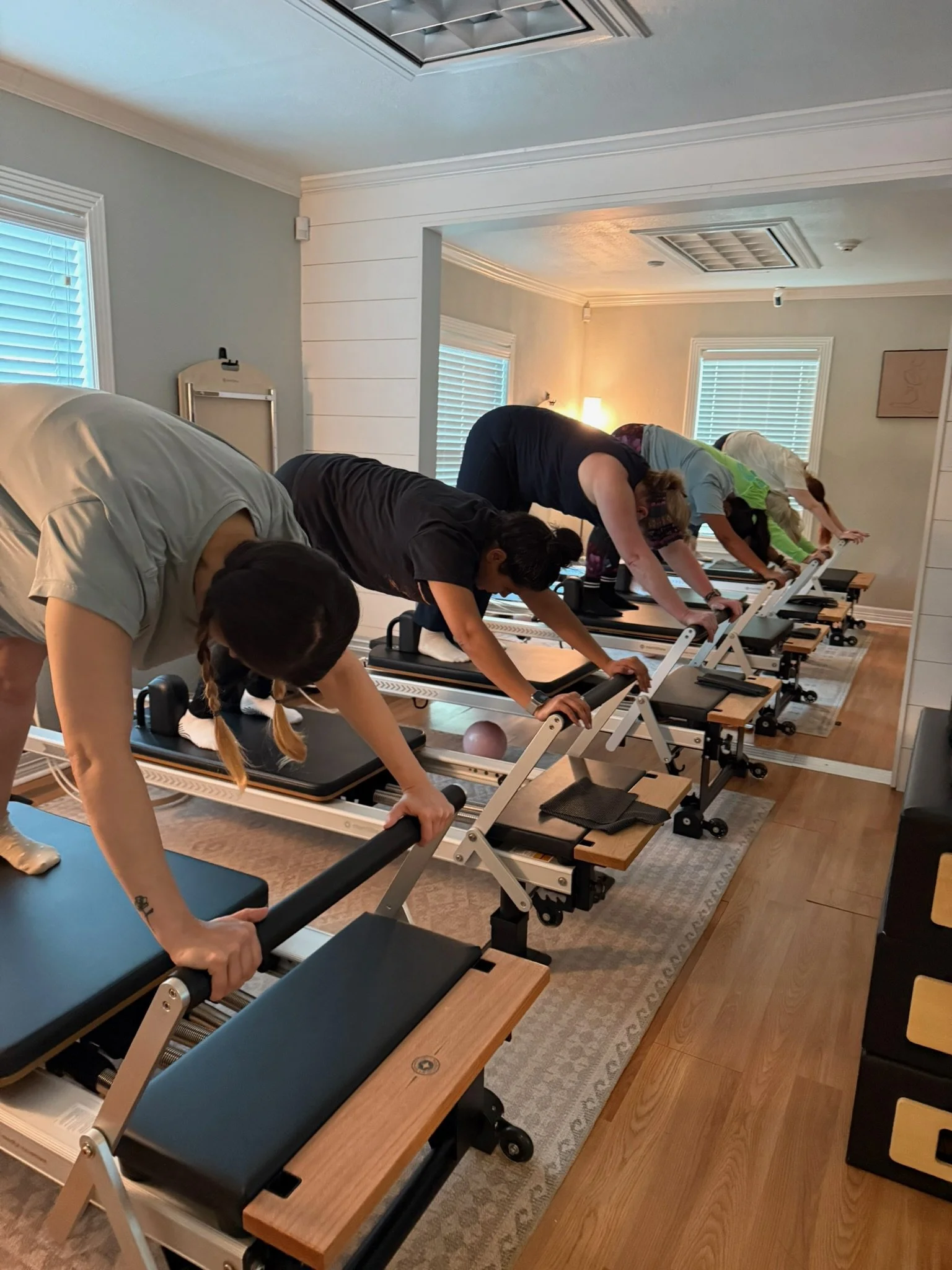 A group of people participating in a Pilates class using reformer machines in a well-lit room with wooden floors and windows with blinds.