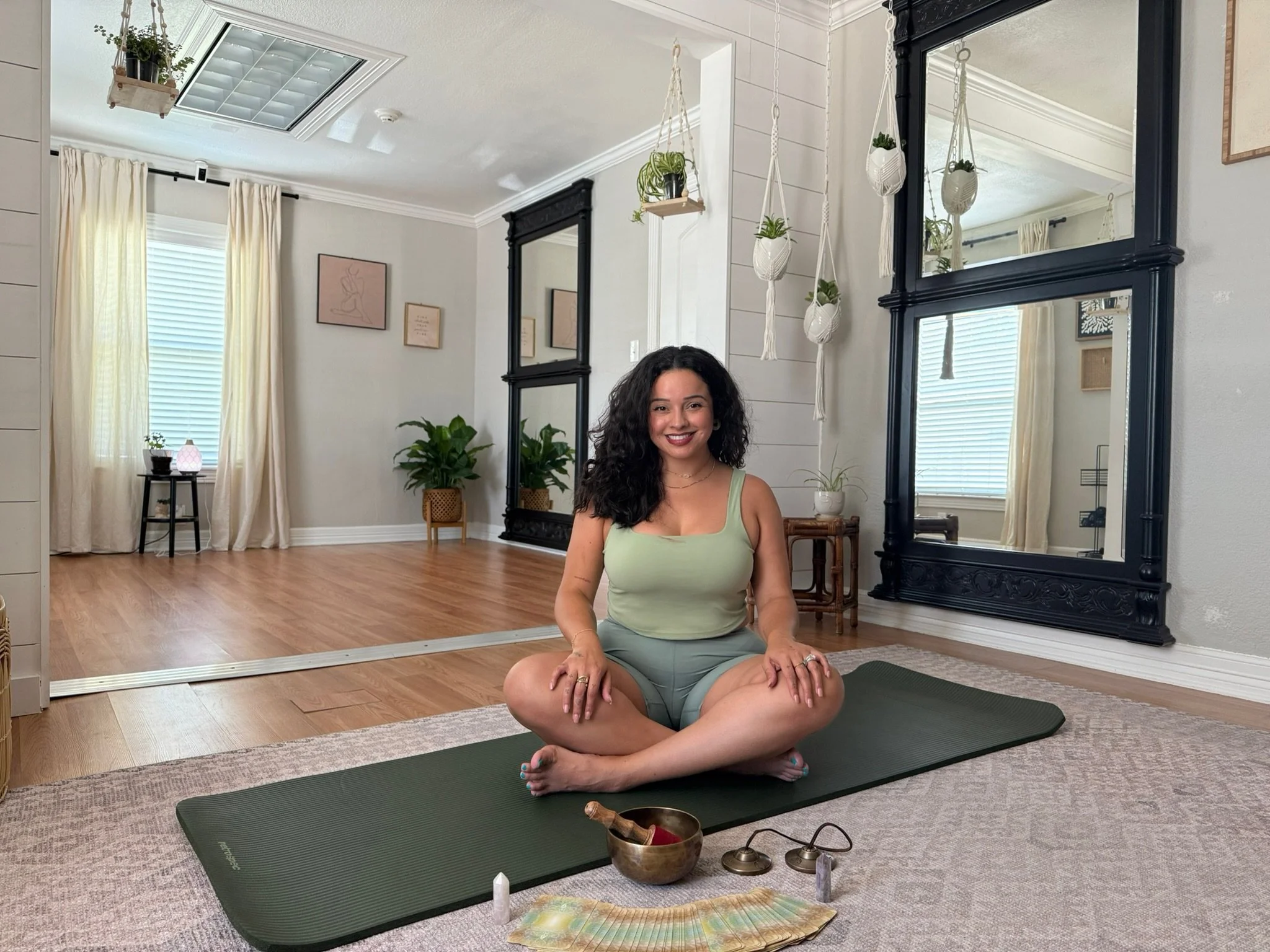 A woman practicing yoga in a bright, cozy living room with plants, framed artwork, and large mirrors. She is seated on a yoga mat in a cross-legged meditation pose, smiling. Yoga accessories and decorative items are visible nearby.