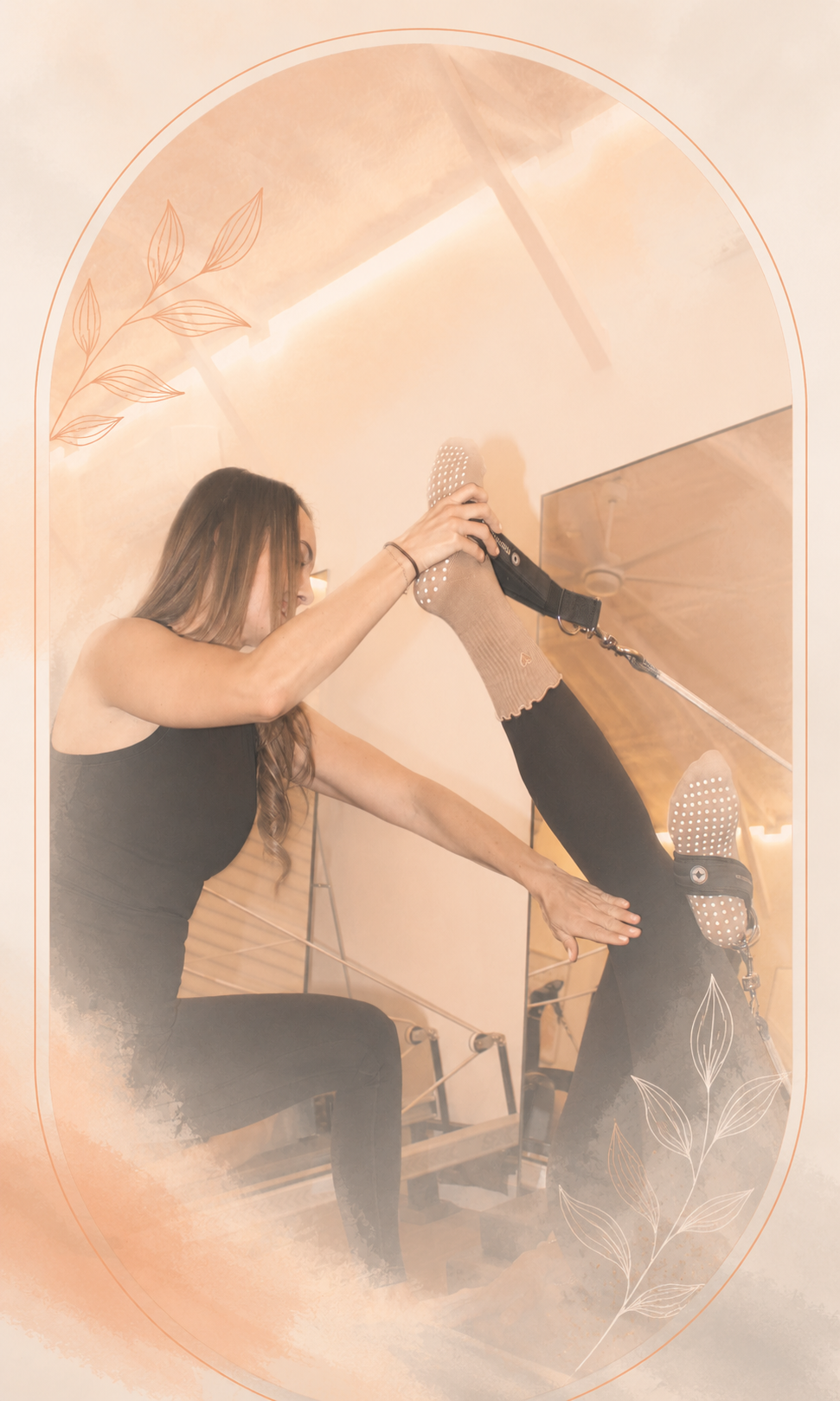 A woman with long hair wearing a black sleeveless top and black leggings is sitting on a Pilates reformer machine in a fitness studio. She is lifting her leg, which is covered with a tan sock, using a strap attached to the machine, while her other hand is placed on her thigh for support.