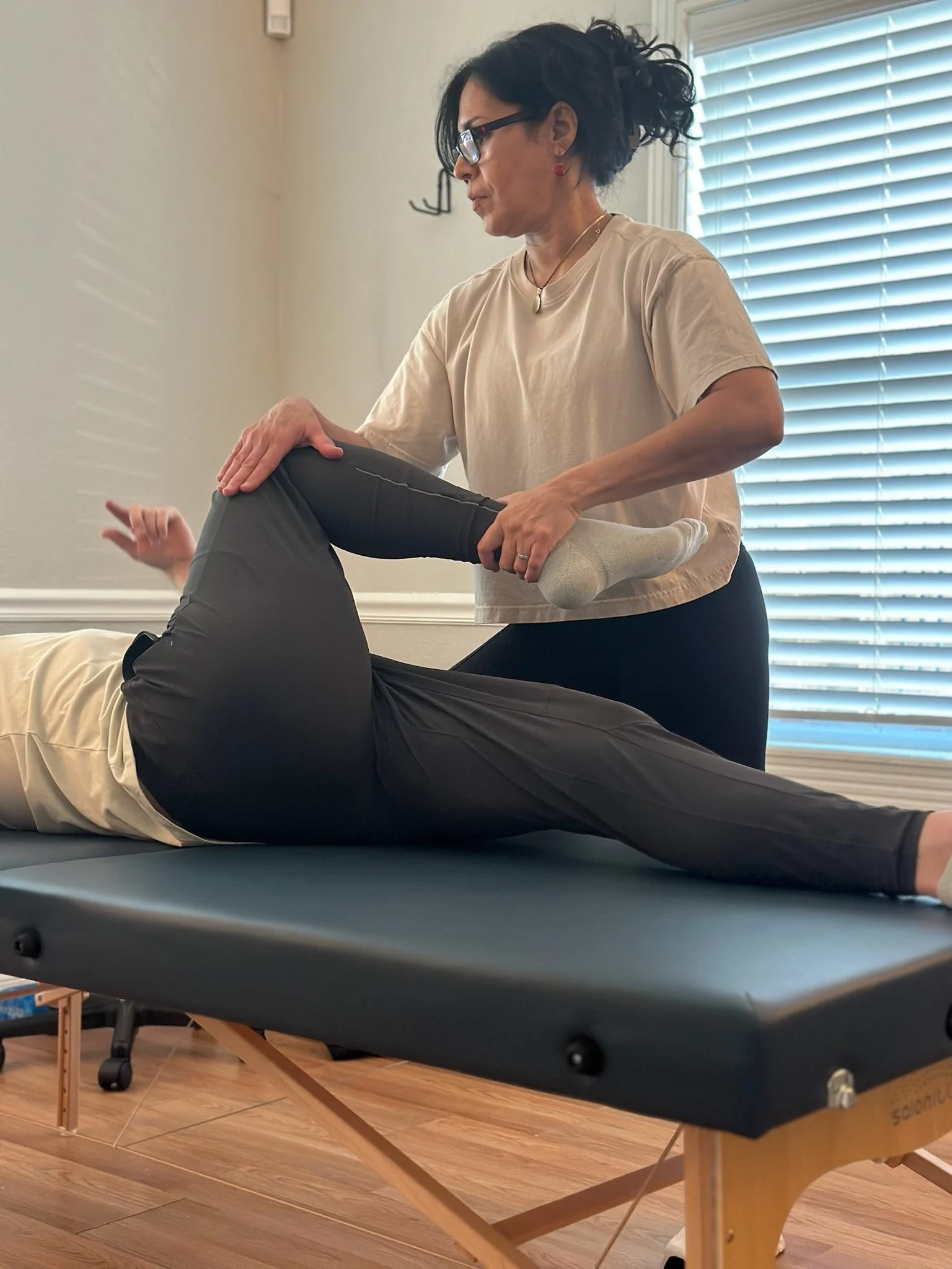 A woman wearing glasses and a beige shirt assists a person performing a leg stretch on a massage table indoors with window blinds in the background.