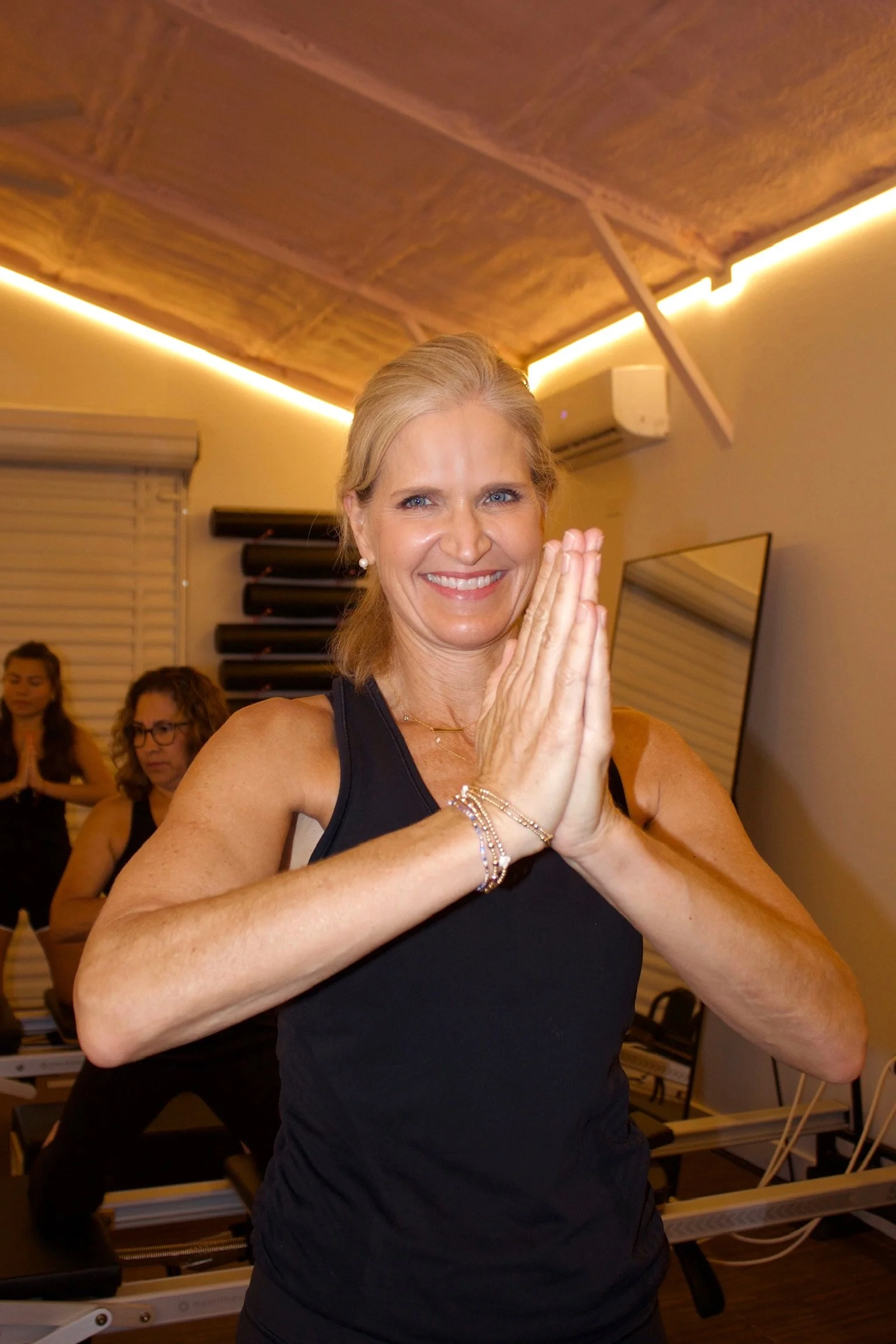 A woman in a black tank top practicing yoga or meditation with her hands pressed together in front of her face in a prayer position, smiling. Several other women are in the background, some also in yoga poses, in a room with wooden ceiling and soft lighting.