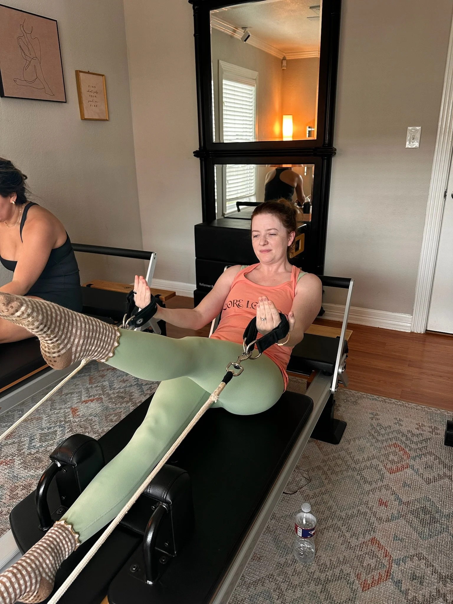 A woman in a yoga pose on a reformer machine, with her leg extended upward and her hands in a meditative position, in a room with a mirror, a water bottle on the floor, and wall decorations.